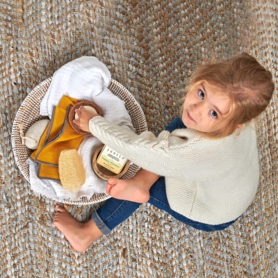 Criança sentada ao lado de cesto com toalha branca, luvas amarelas, escova natural e frasco