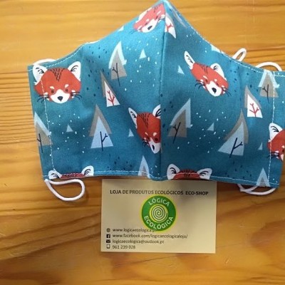 Máscara de tecido azul com estampa de raposas e pinheiros sobre mesa de madeira, com cartão de loja ecológica.