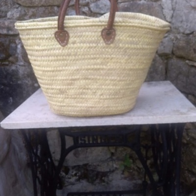 Sacola de palha com pegas de couro sobre mesa de ferro com tampo branco frente a parede de pedra