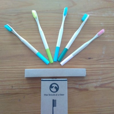 Cinco escovas de dentes com cabos brancos e cores diversas, caixa de papelão com frase 'One brush at a time', sobre superfície de madeira.