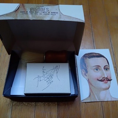 Caixa aberta com pincel, bloco de papel e cartão com ilustração de homem com bigode sobre superfície de madeira.