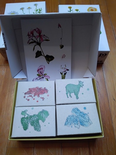 Caixa aberta com quatro sabonetes ilustrados e imagem floral no interior da tampa.