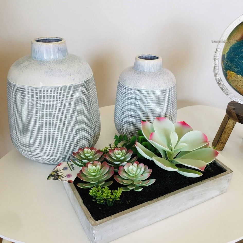Dois vasos cerâmicos e arranjo de suculentas artificiais sobre mesa branca
