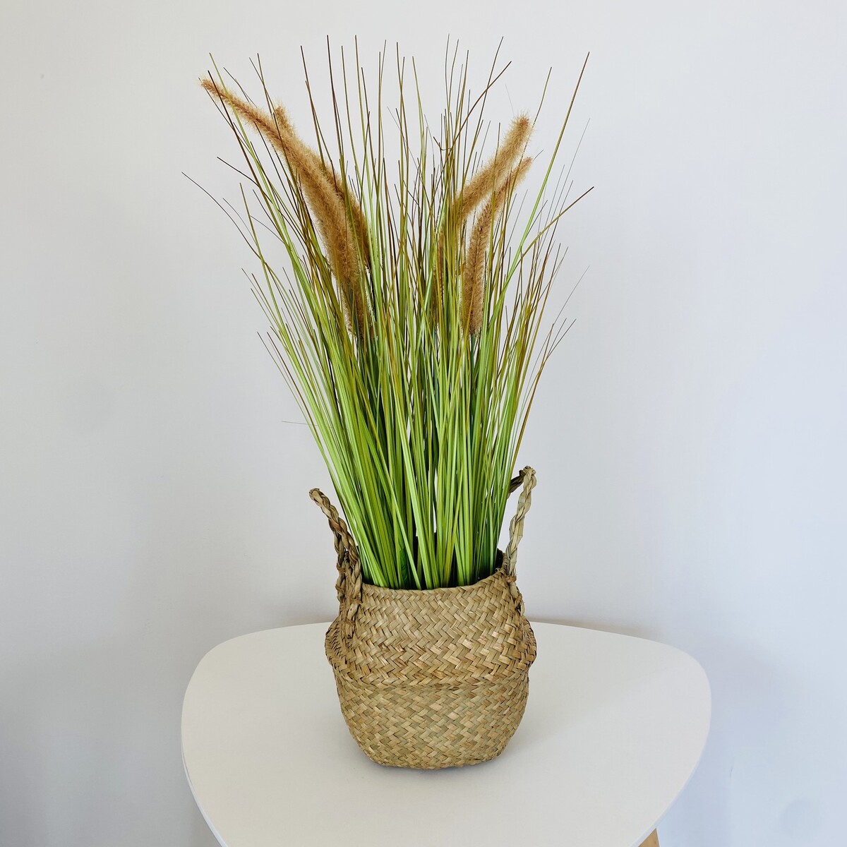 Cesta trançada natural com plantas artificiais verdes e espigas castanhas sobre mesa branca
