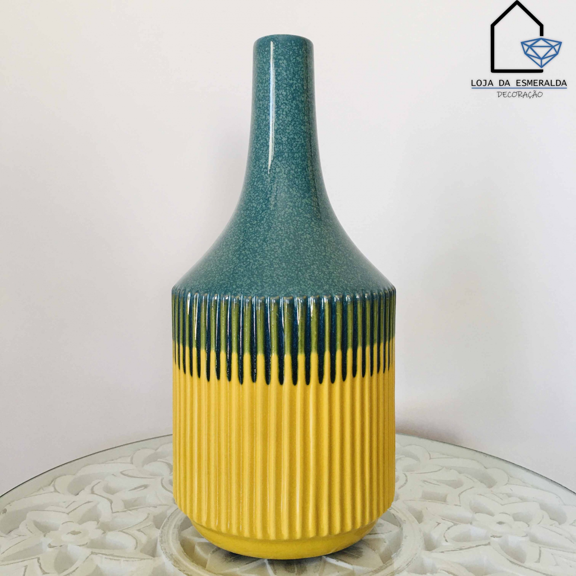 Vaso decorativo cerâmico verde azulado e amarelo sobre mesa branca com padrão floral.