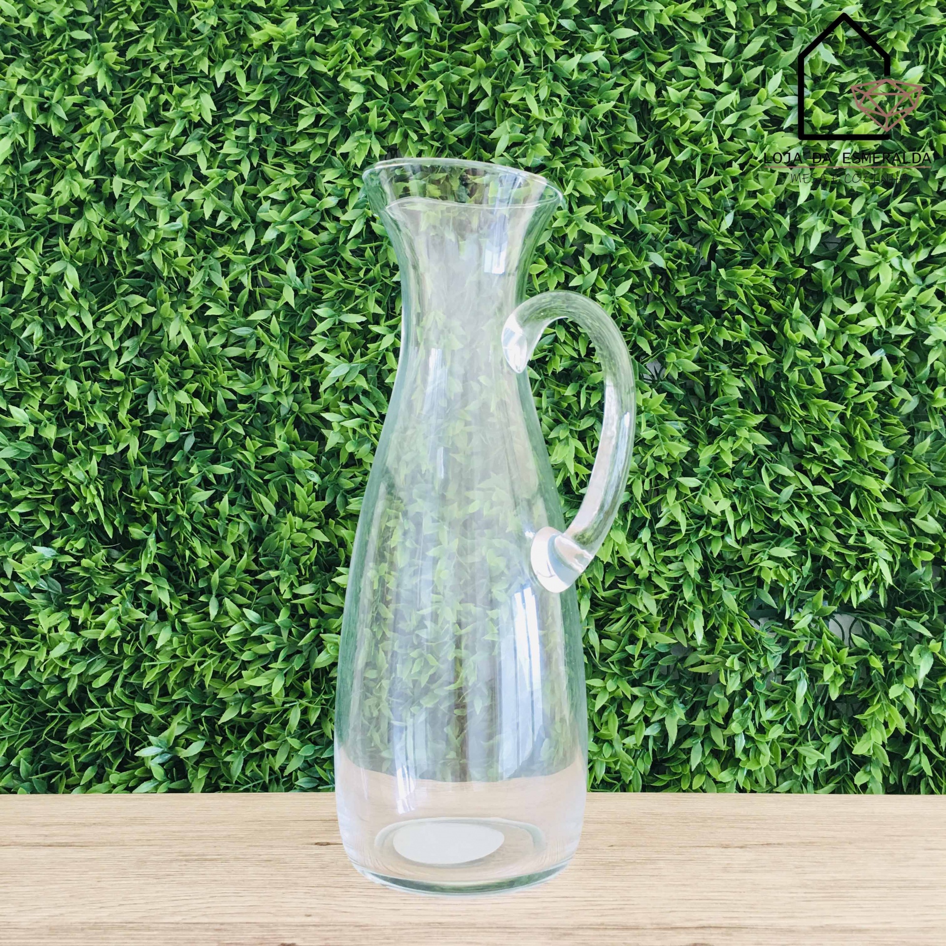 Jarra de vidro transparente com pega sobre mesa de madeira, fundo de folhas verdes