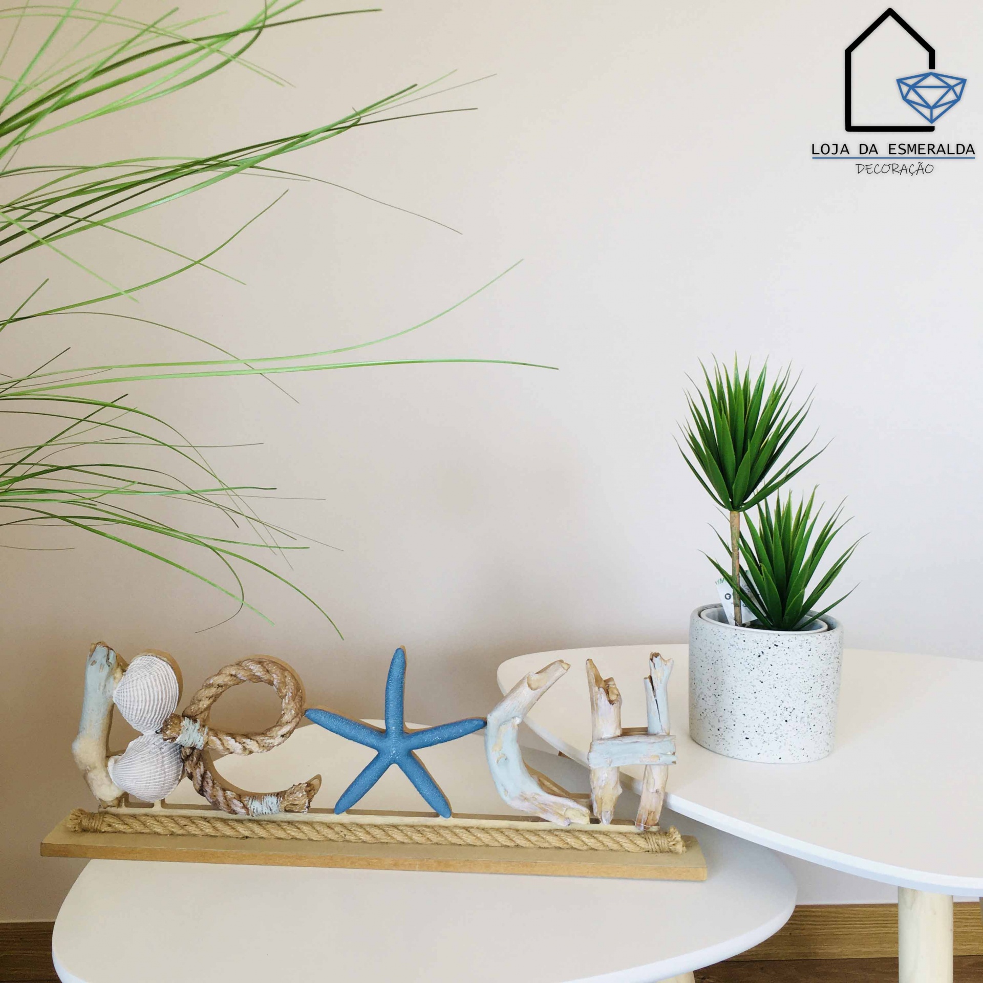 Decoração de mesa com palavra LOVE em madeira, corda, conchas e estrela do mar azul, ao lado de planta em vaso cinza sobre mesas brancas.
