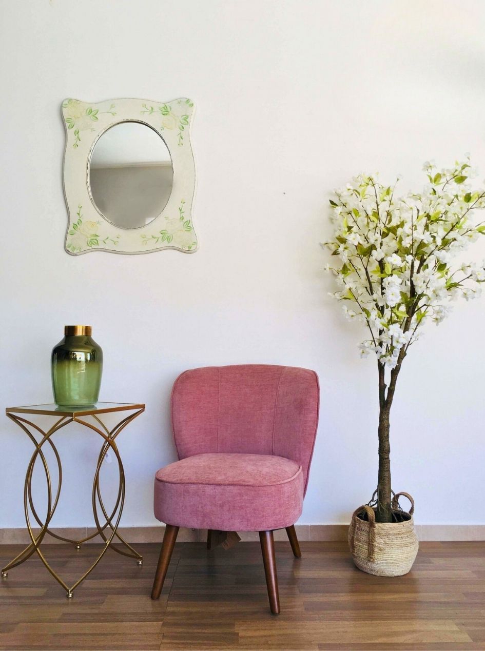 Cadeira rosa, mesa metálica dourada com jarro verde, espelho oval com moldura floral e árvore artificial em vaso de fibra.