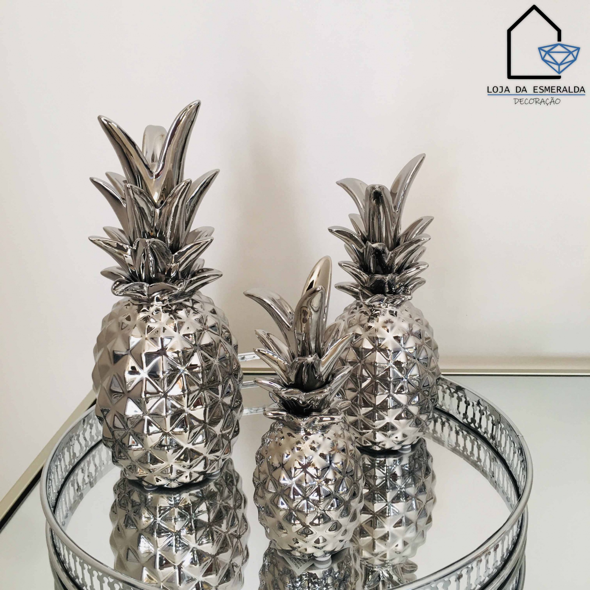 Três abacaxis decorativos prateados sobre bandeja espelhada e logotipo na parede