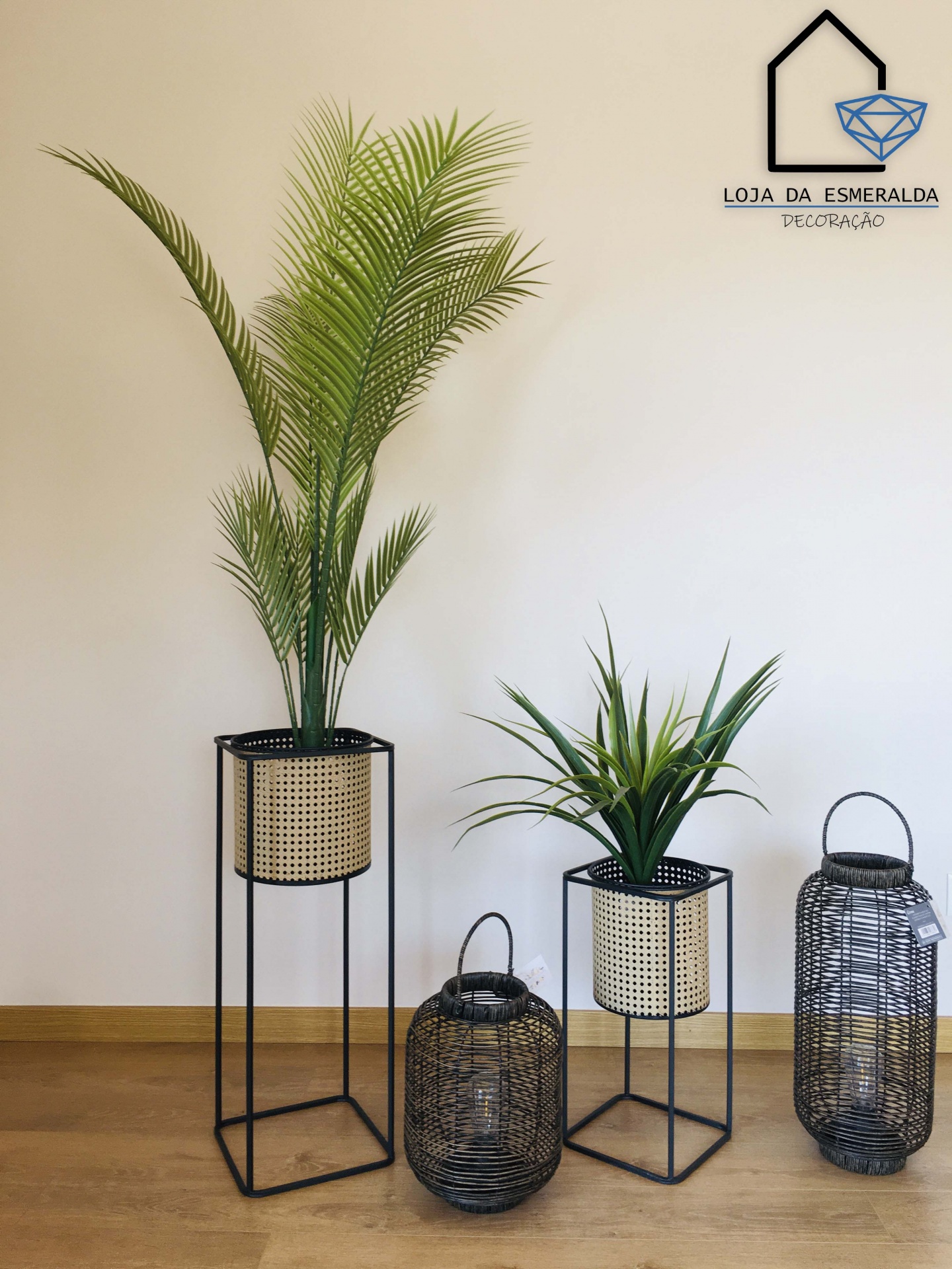 Duas plantas em vasos de ráfia com suporte metálico, lanternas de metal preto, sobre chão de madeira clara, parede branca com texto e logo da loja.