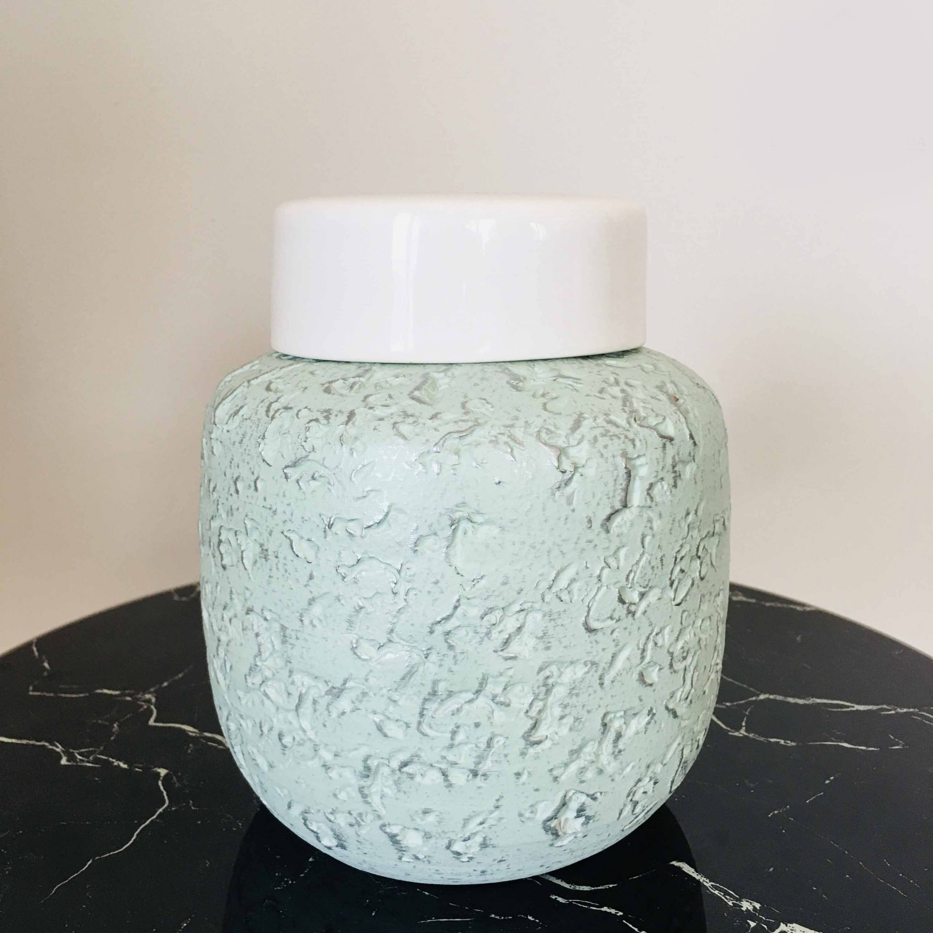 Jarro decorativo verde água com tampa branca sobre superfície preta com padrão branco