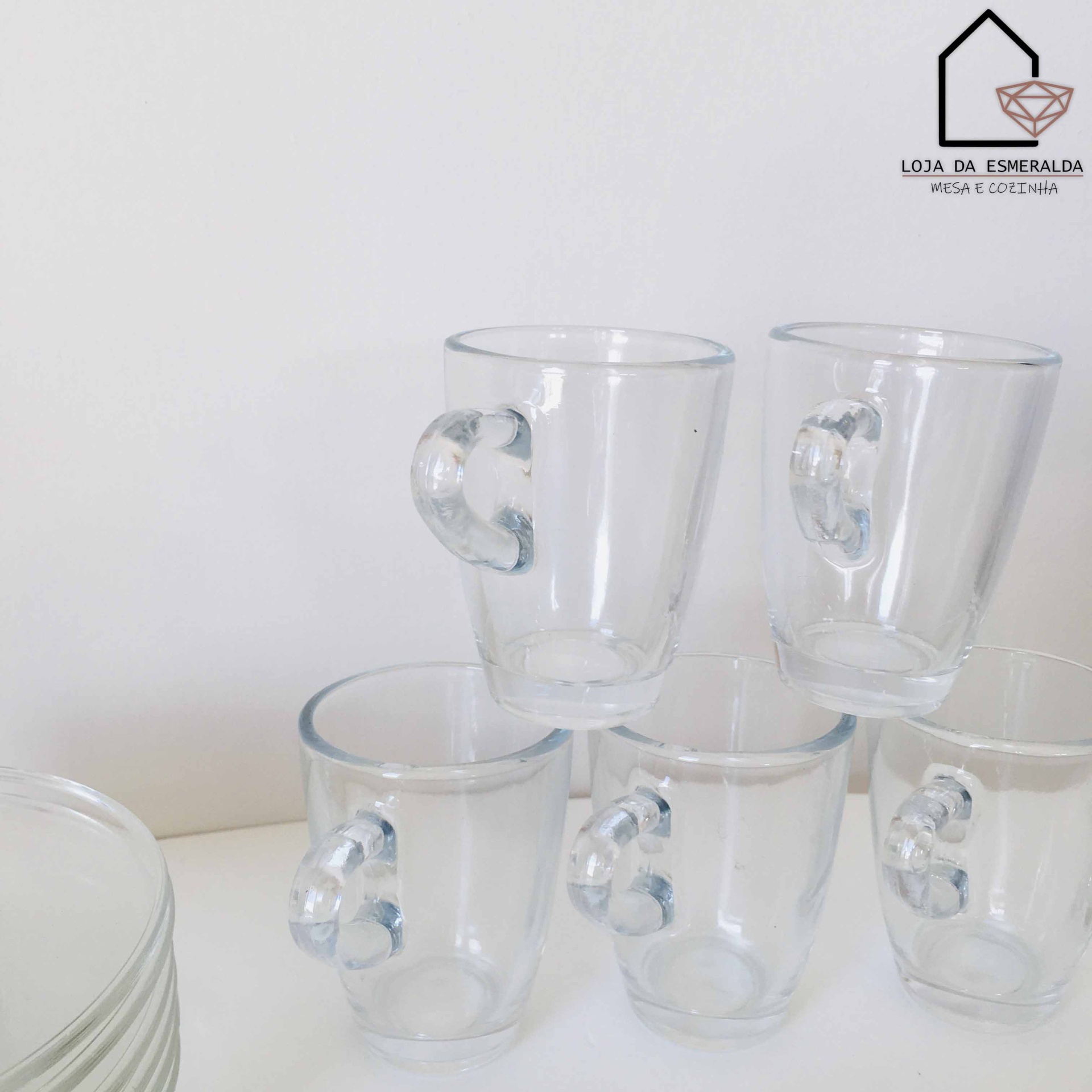 Seis copos de vidro com pega em forma de fita sobre superfície branca e pratos empilhados