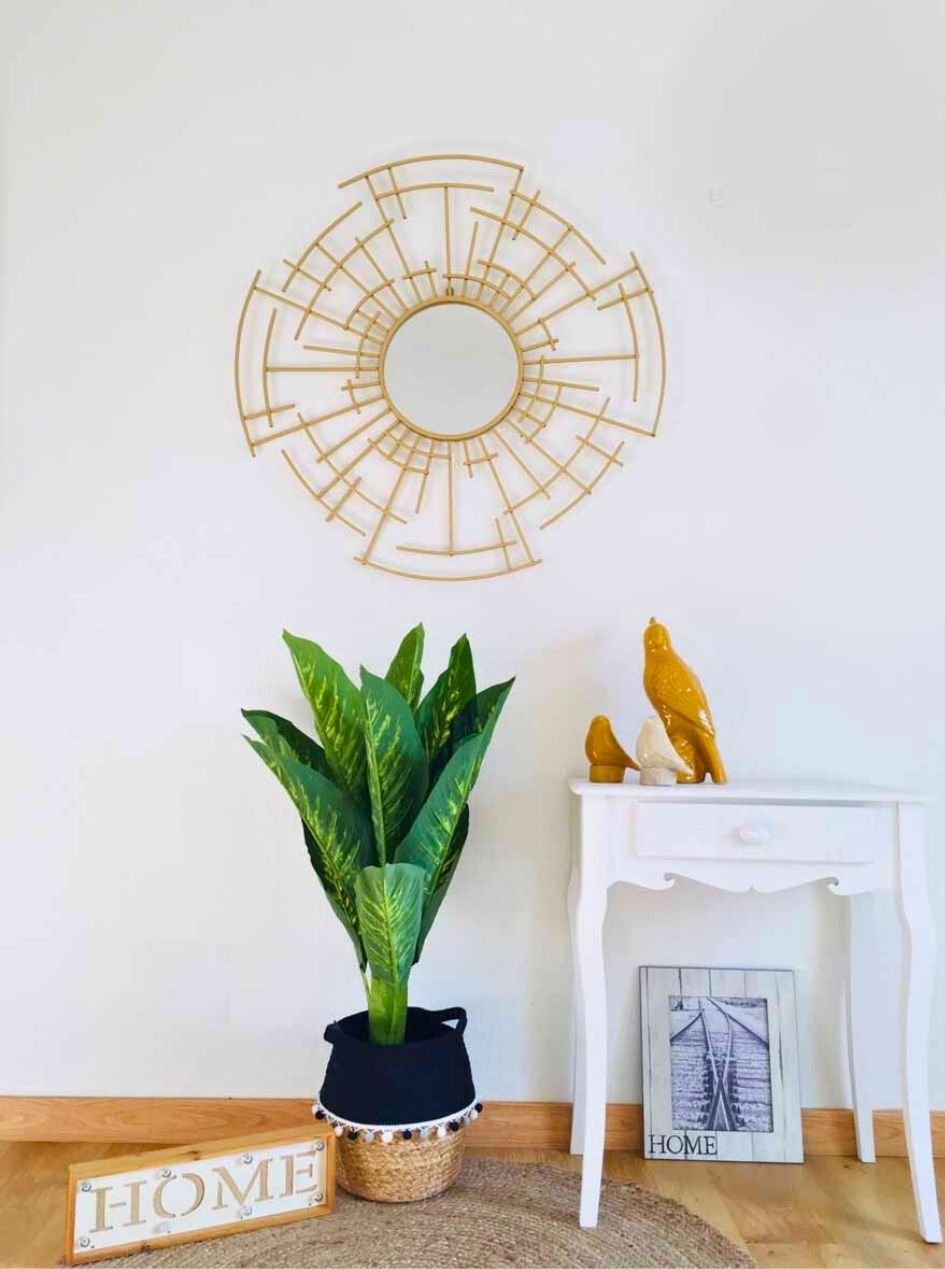 Espelho decorativo dourado, planta em vaso, mesa branca com esculturas e elementos decorativos em ambiente interior.