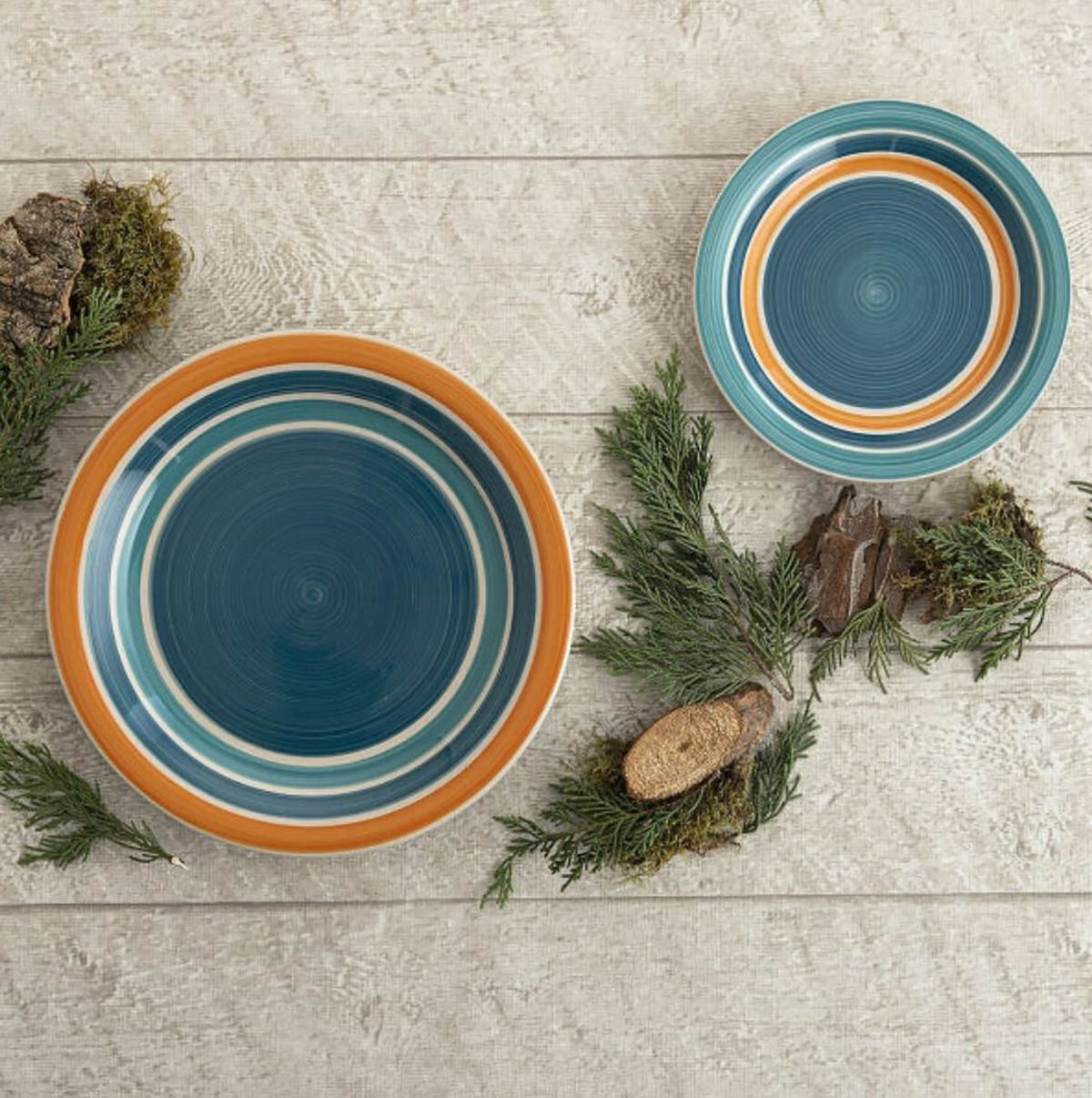 Dois pratos coloridos redondos com motivos de anéis em verde-azulado, branco e laranja sobre superfície de madeira com decoração natural