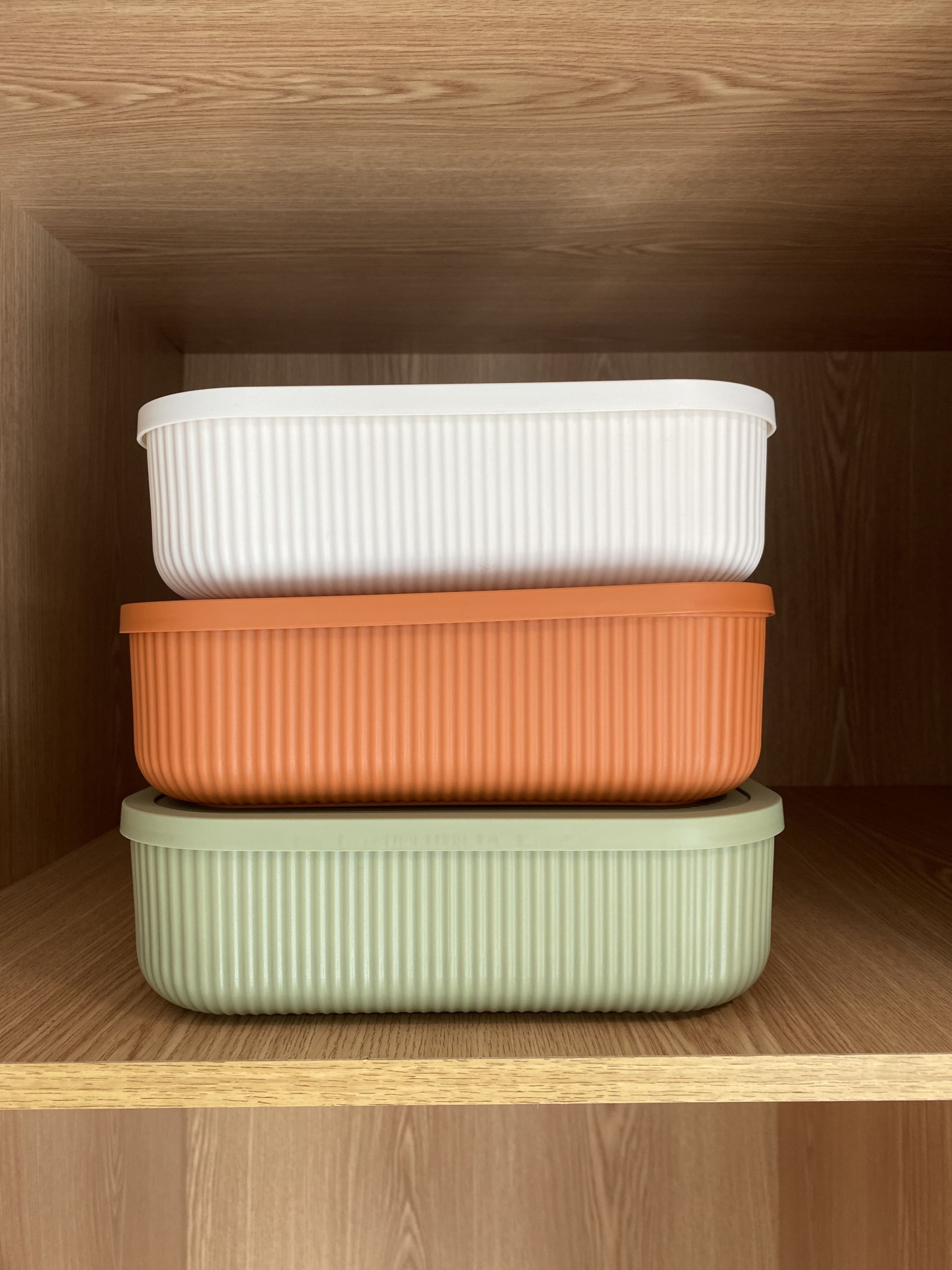Três caixas retangulares plástico branco, laranja e verde empilhadas em prateleira de madeira