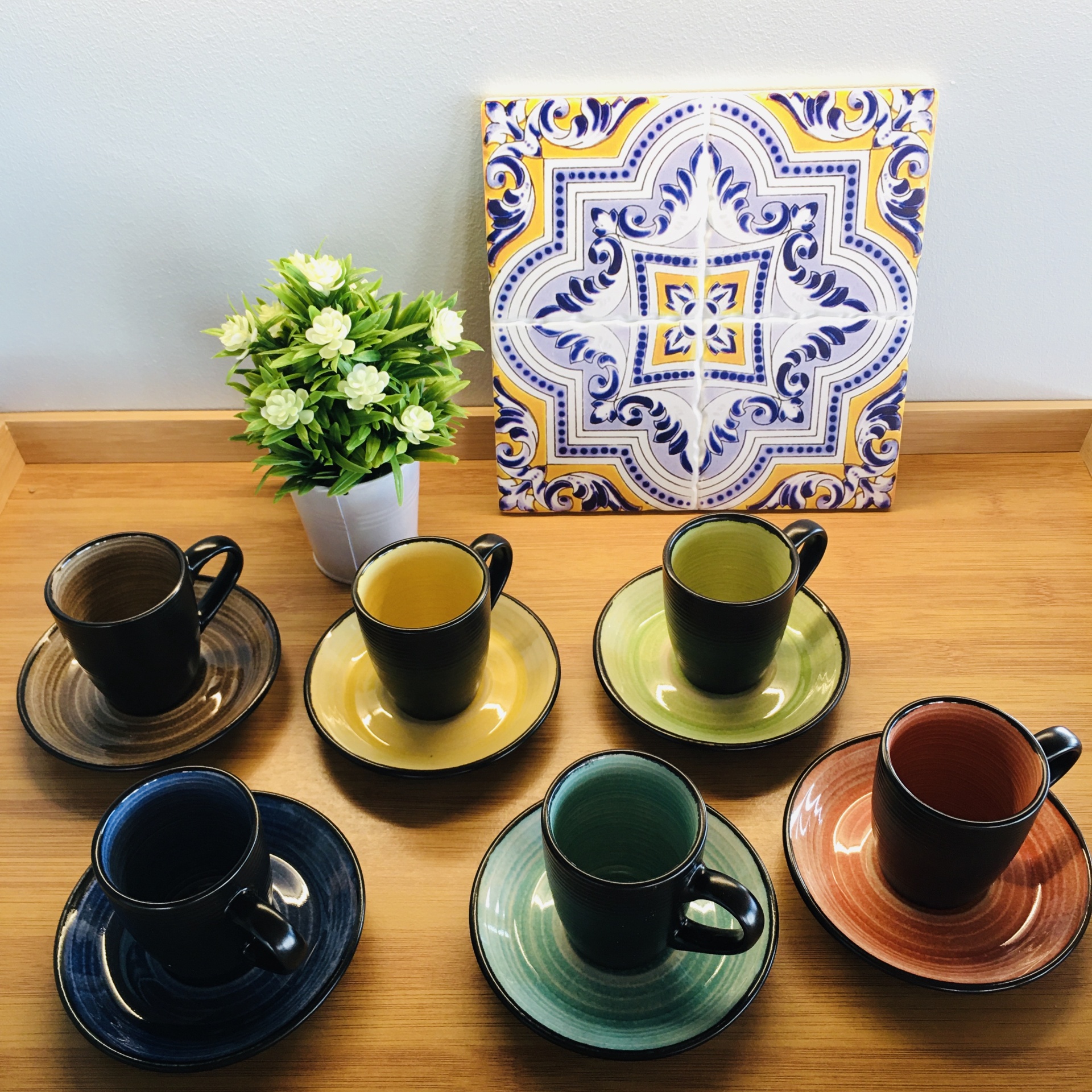 Seis chávenas de chá coloridas com pires, planta artificial e azulejo decorativo atrás.