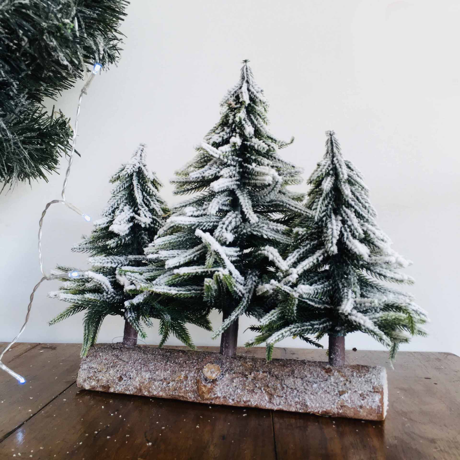 Três mini árvores de Natal artificiais com neve sobre base de tronco em madeira