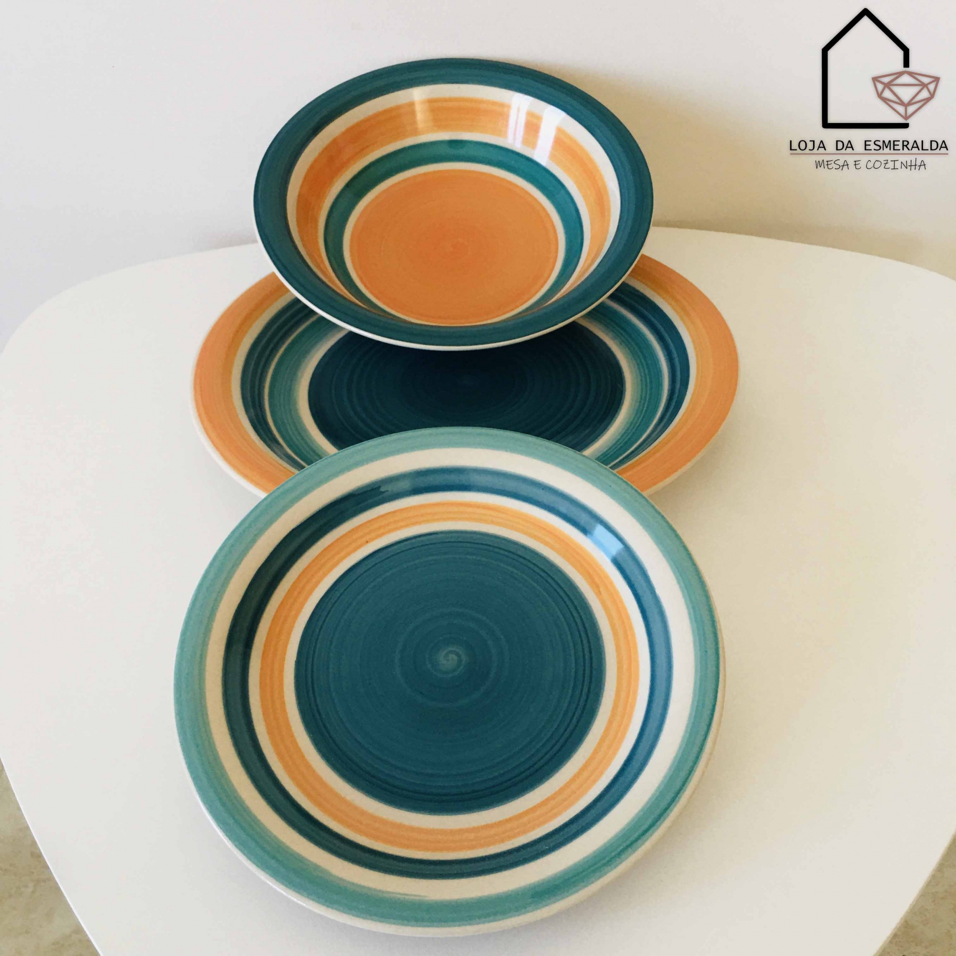 Três pratos coloridos com padrão circular em azul, castanho e branco sobre mesa branca, ao lado logotipo da loja.