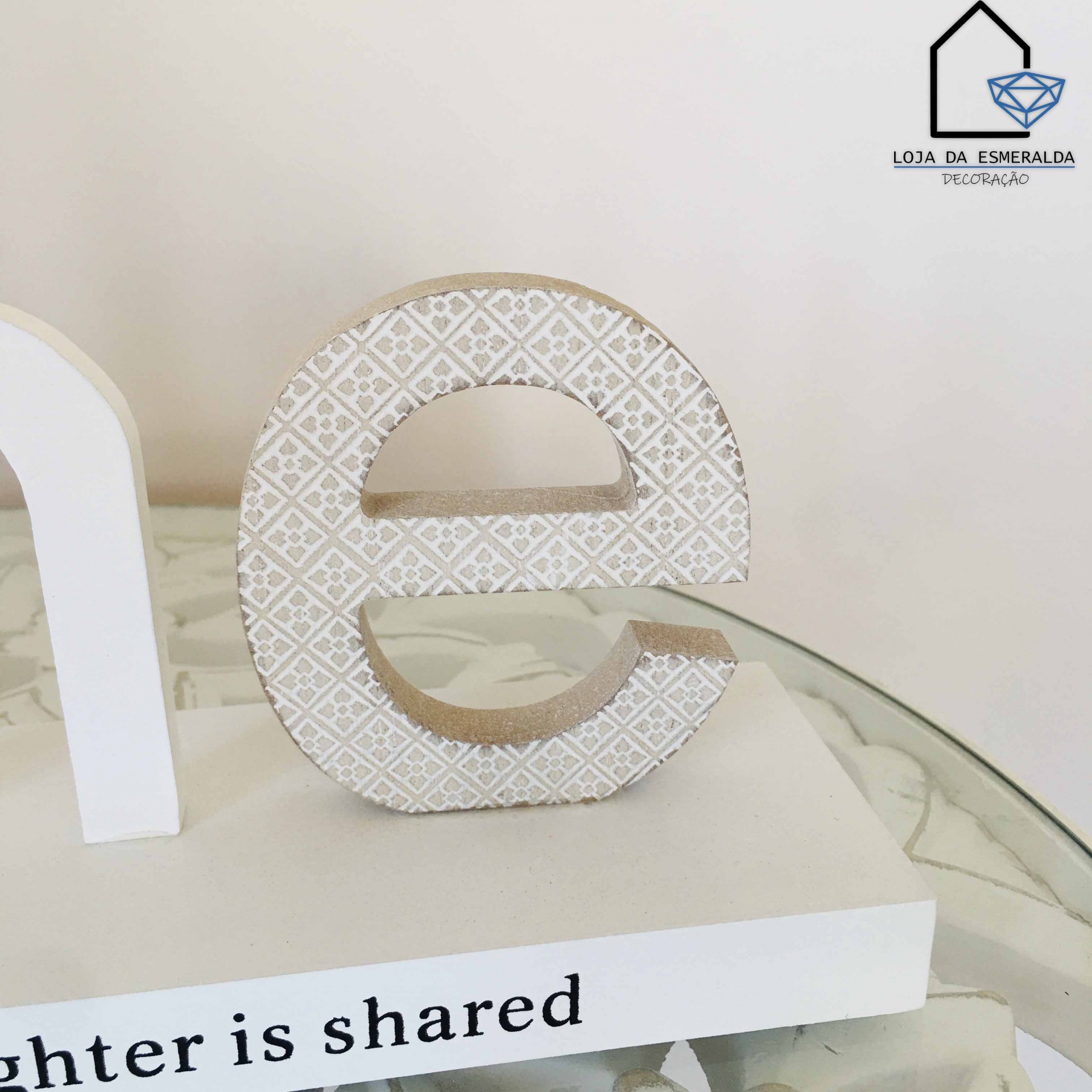 Letra decorativa minúscula 'e' em madeira com padrão branco e bege sobre superfície branca com texto parcial visível