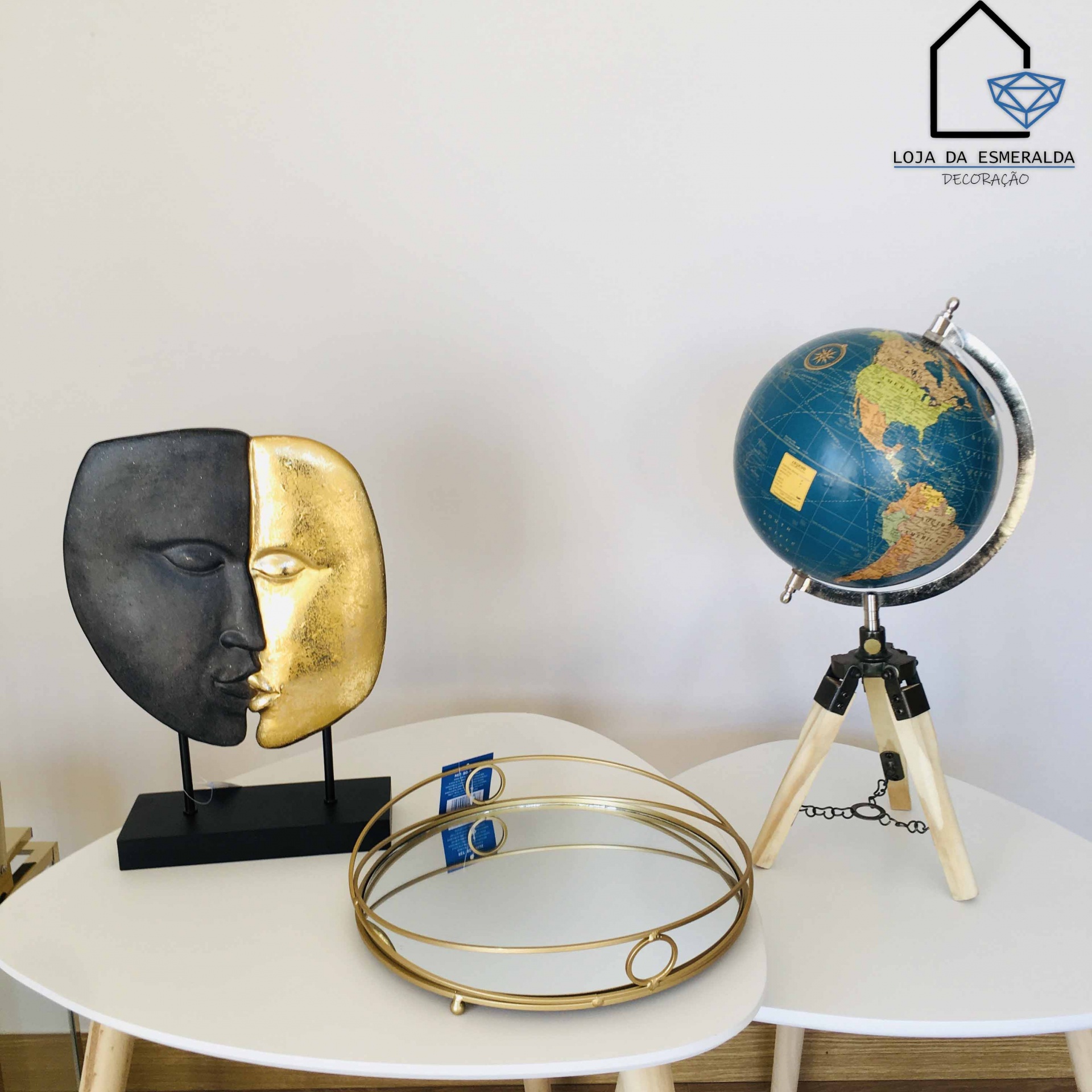 Máscara decorativa metade preta metade dourada, globo terrestre em tripé de madeira clara e bandeja redonda dourada com espelho em mesas brancas