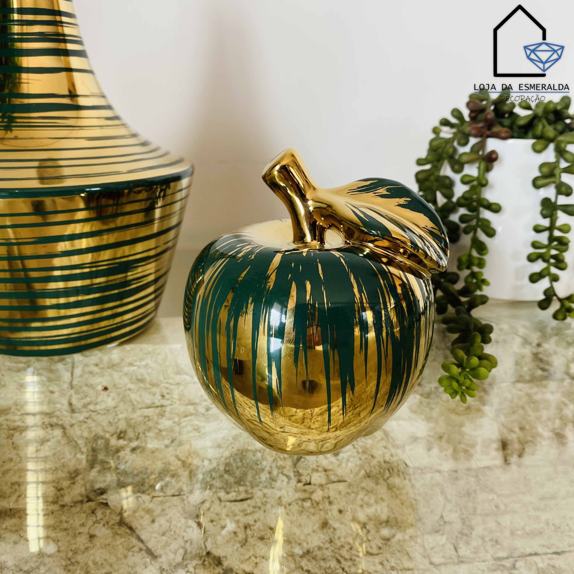 Objeto decorativo maçã dourada e verde com vaso e planta ao fundo sobre superfície marmoreada
