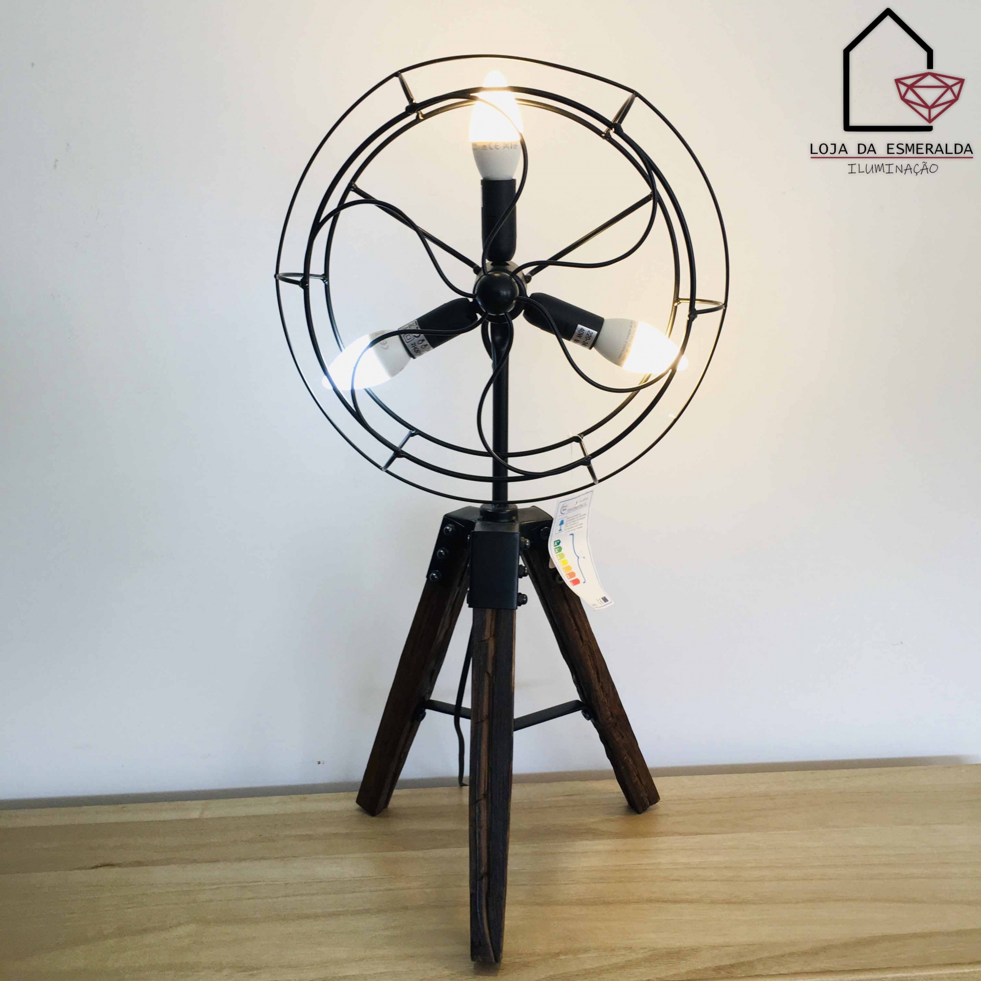 Candeeiro de mesa tripé em madeira escura e metal preto com três lâmpadas ligado sobre mesa de madeira clara