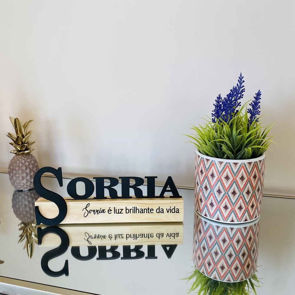 Placa decorativa com a palavra SORRIA e vaso com plantas artificiais ao lado sobre mesa espelhada.