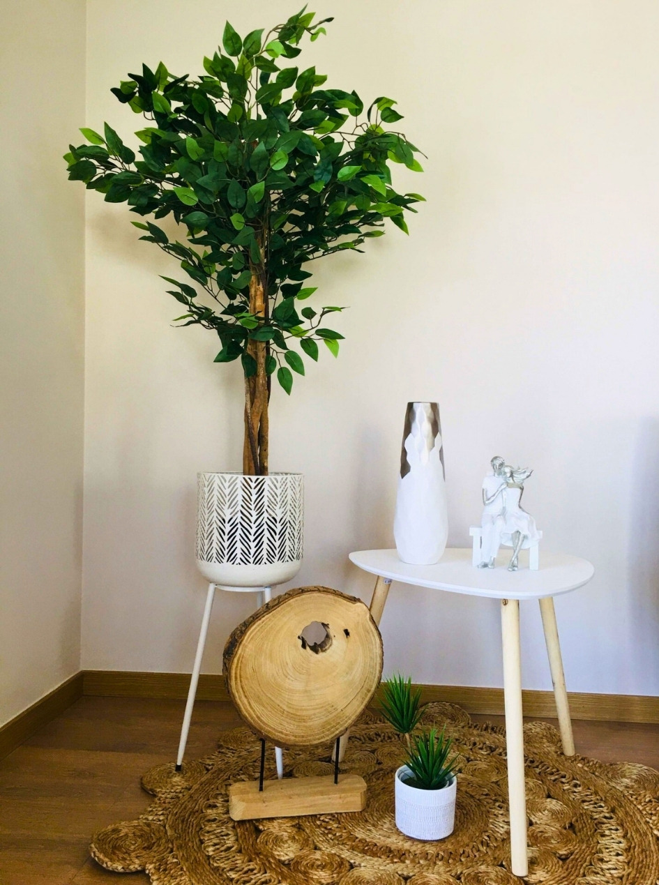 Planta artificial em vaso branco, mesa branca com vaso e escultura, tronco decorativo e tapete amarelo trançado.