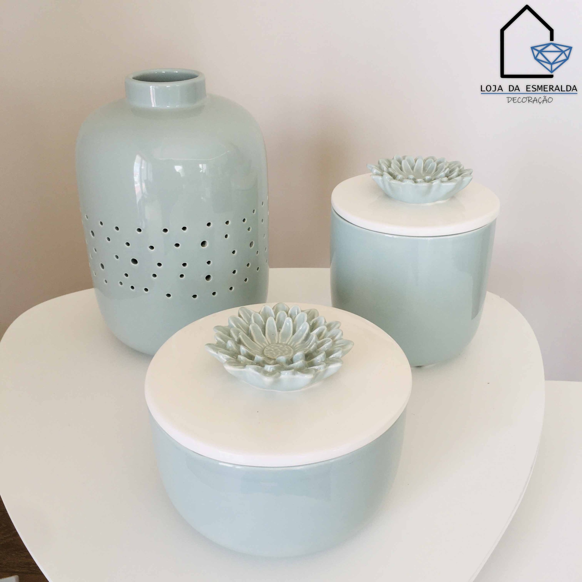 Conjunto de cerâmica verde clara com vaso perfurado e potes com flores decorativas em tampas brancas sobre mesa branca
