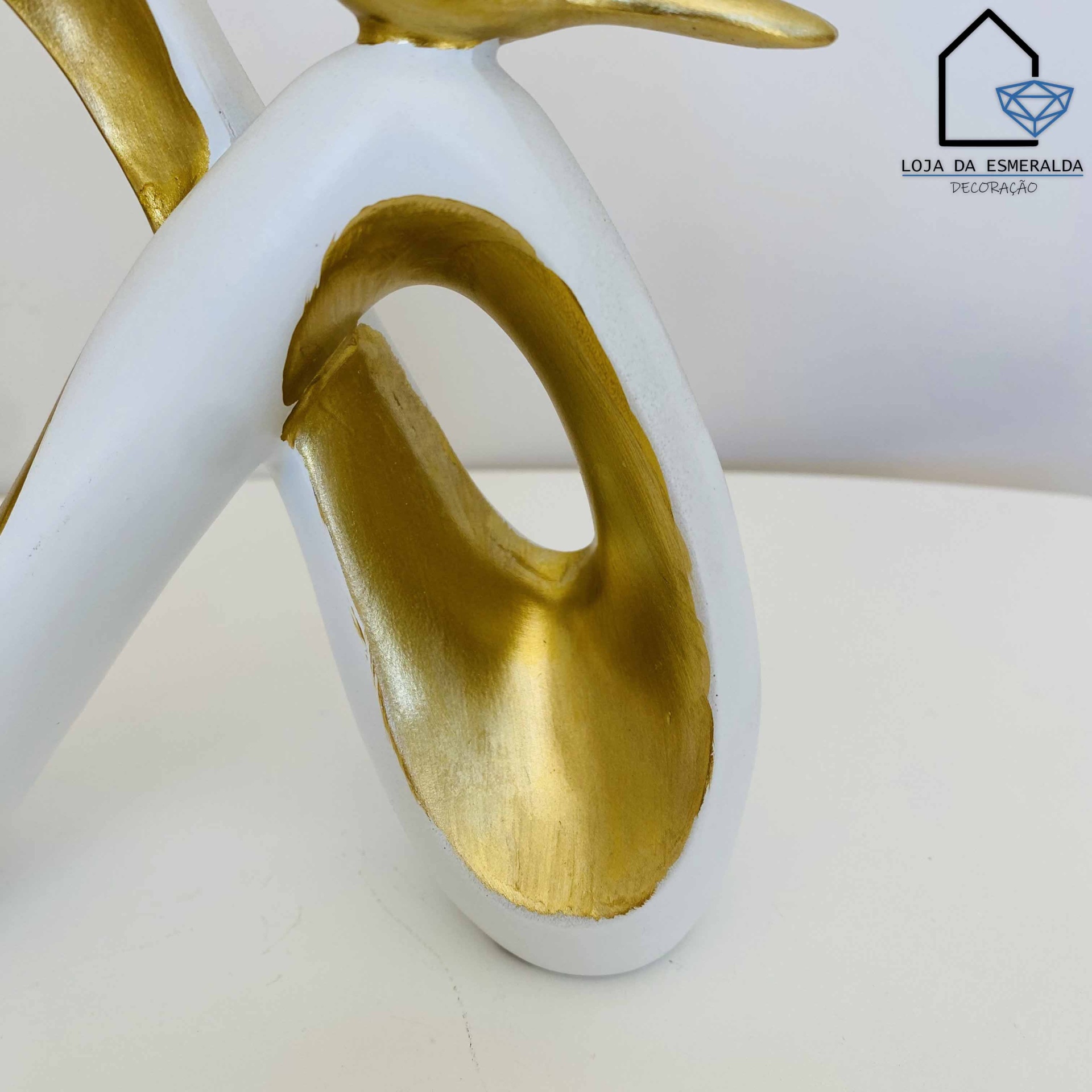 Escultura decorativa branca e dourada abstrata em superfície branca com logotipo de loja no fundo.