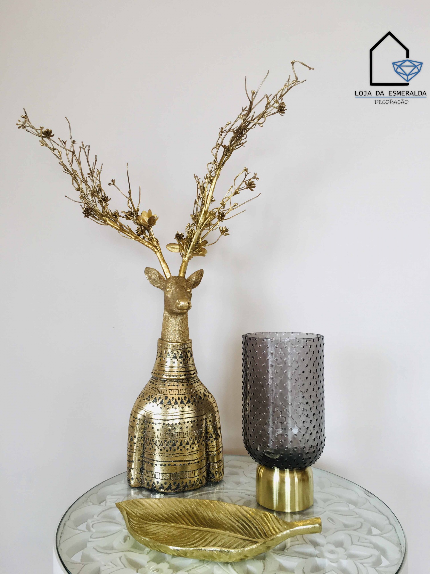 Decoração com cervo dourado, vaso cinzento texturizado e prato folha dourada numa mesa redonda de vidro.