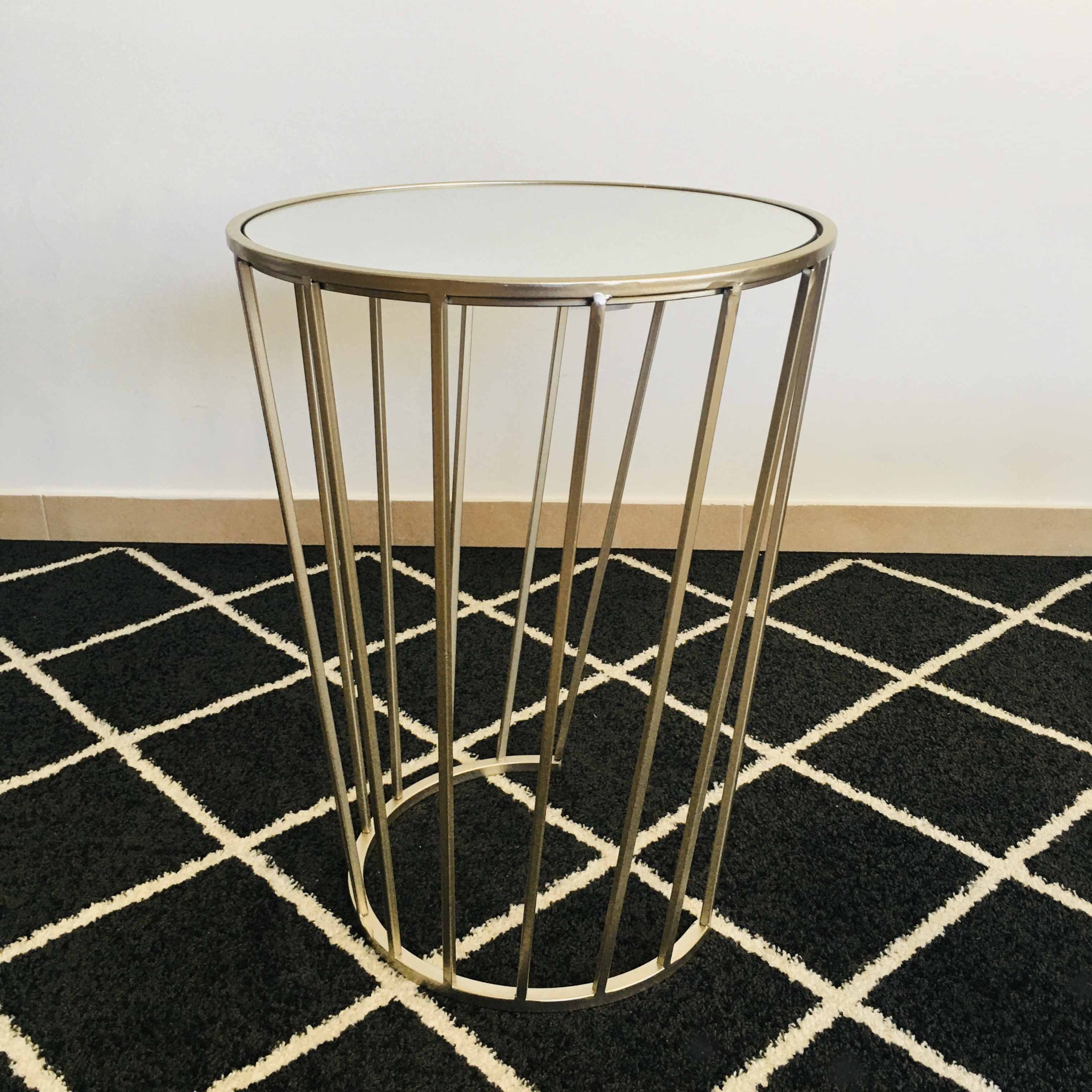 Mesa lateral redonda com tampo branco e base metálica dourada sobre tapete preto com losangos brancos