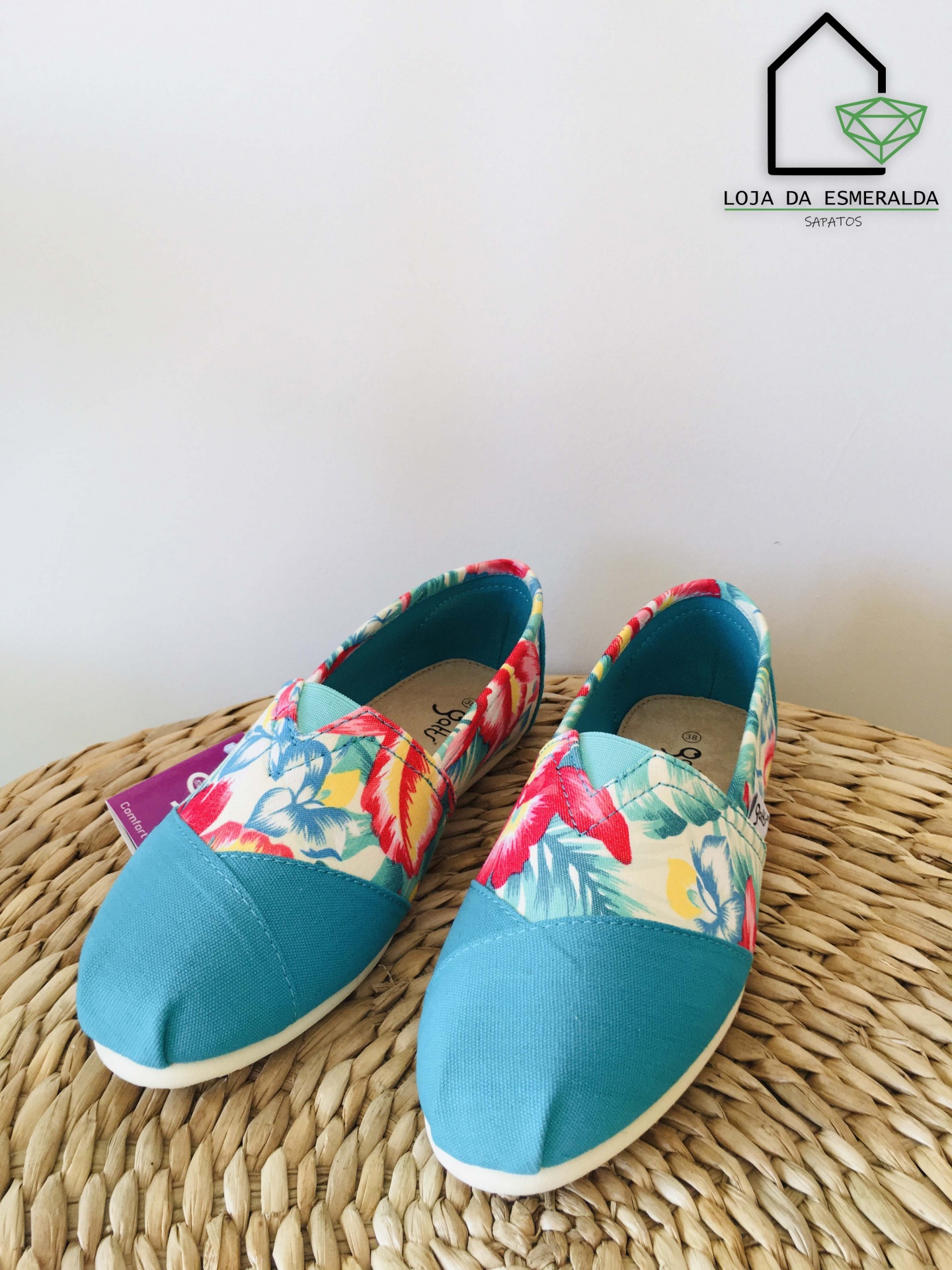 Sapatos alpargatas turquesa e floral sobre superfície de corda com logo ao fundo