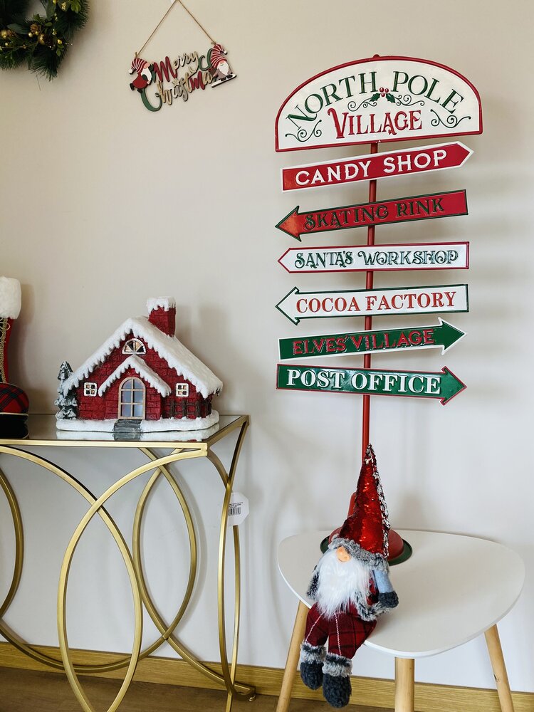 Placa decorativa do tipo indicativo de madeira com temas de Natal e um boneco de duende pendurado numa mesa branca junto a uma casa decorativa