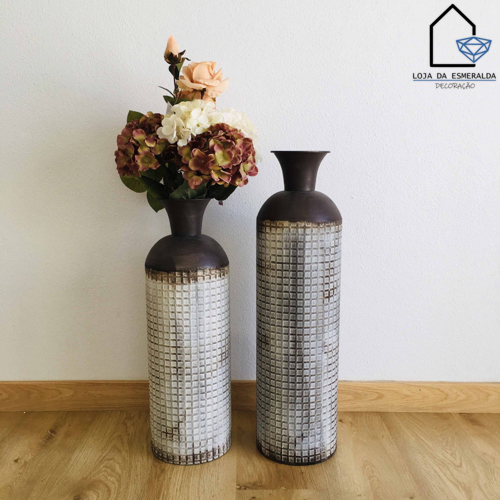 Dois vasos decorativos altos com textura quadriculada, um com flores artificiais, sobre chão de madeira e parede branca