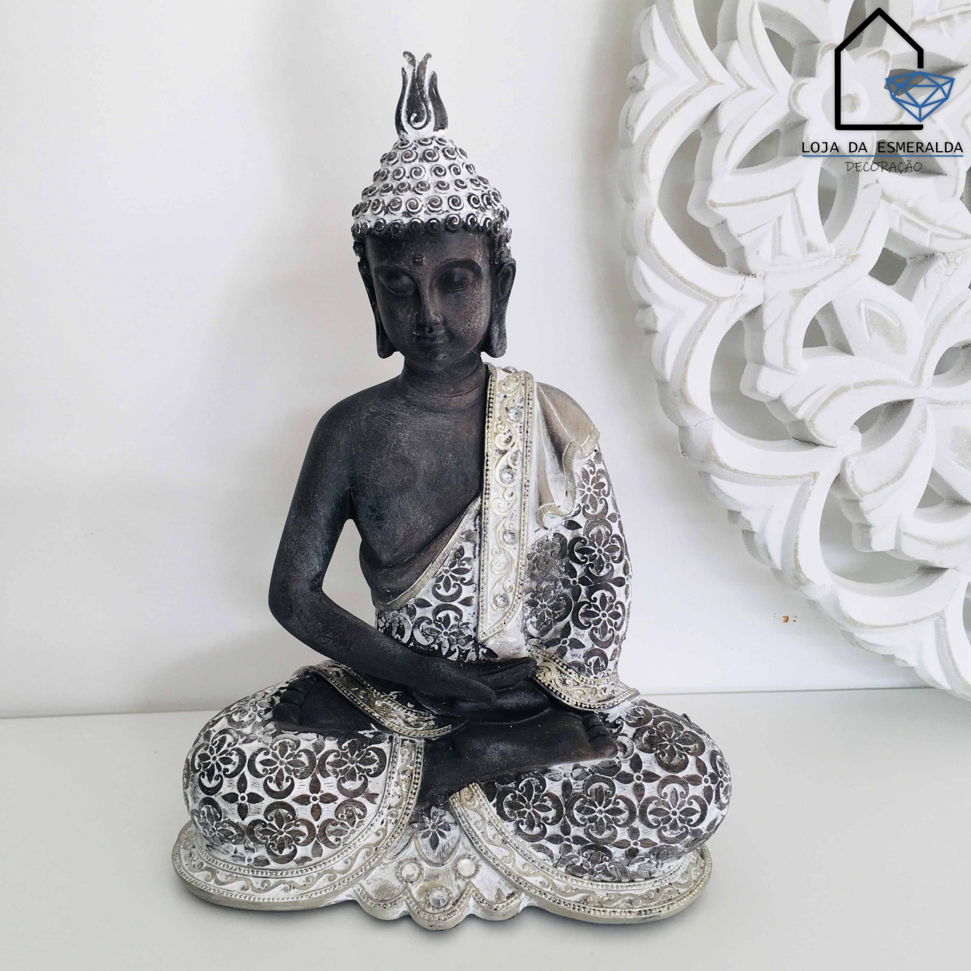 Estátua de Buda decorativa com manto branco e prateado em padrão floral, sobre base branca com fundo claro e decoração branca em relevo.