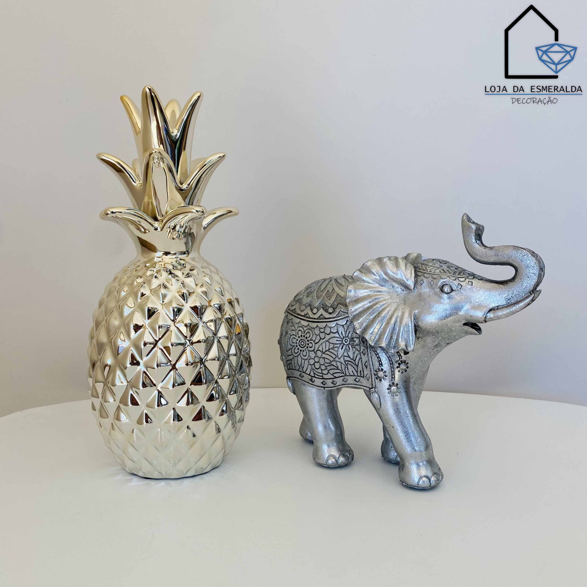 Peças decorativas abacaxi dourado e elefante prateado com detalhes em padrão tribal sobre superfície branca.