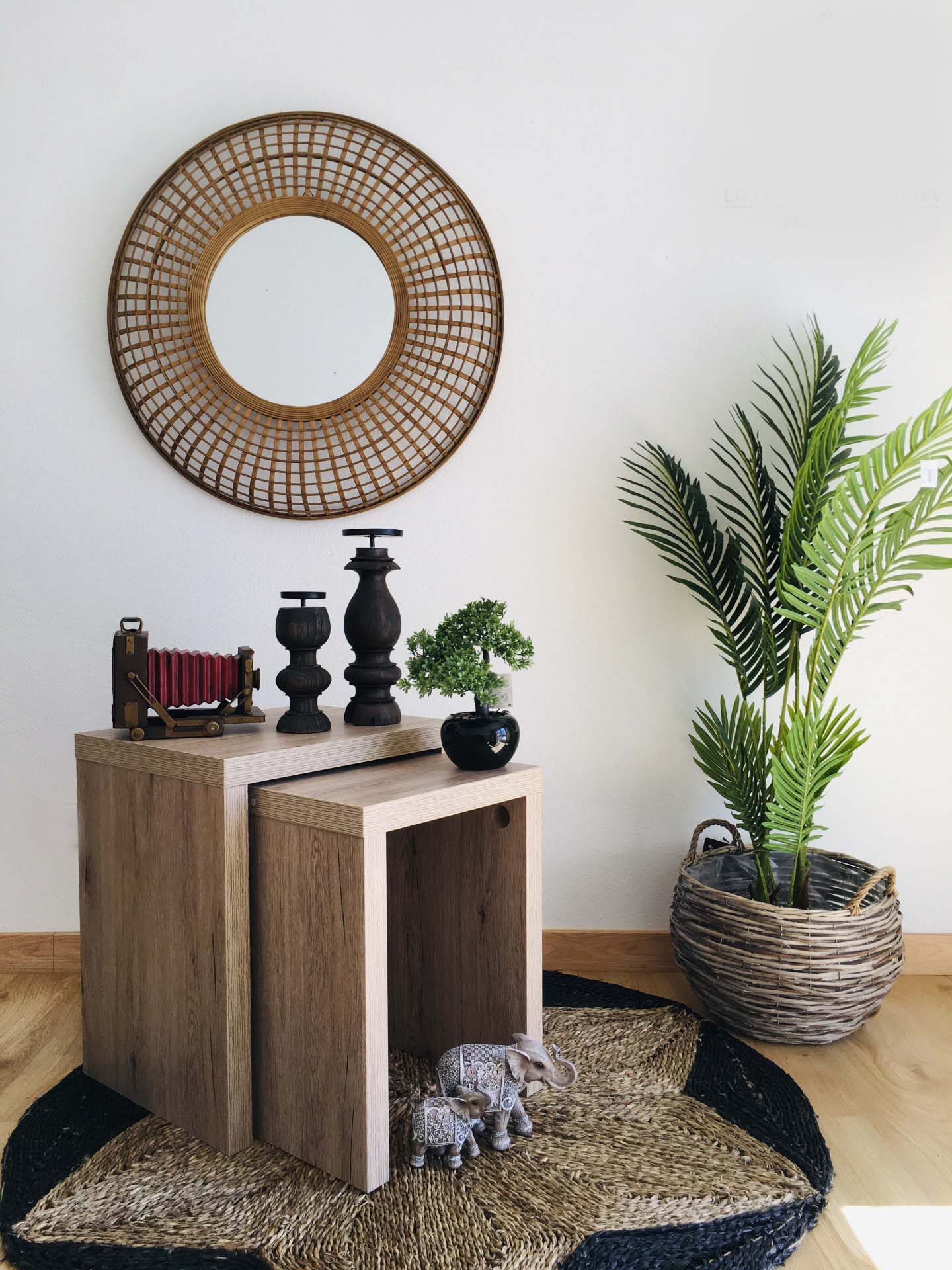 Espaço decorativo com mesas de madeira, espelho redondo de rattan, plantas e objetos decorativos