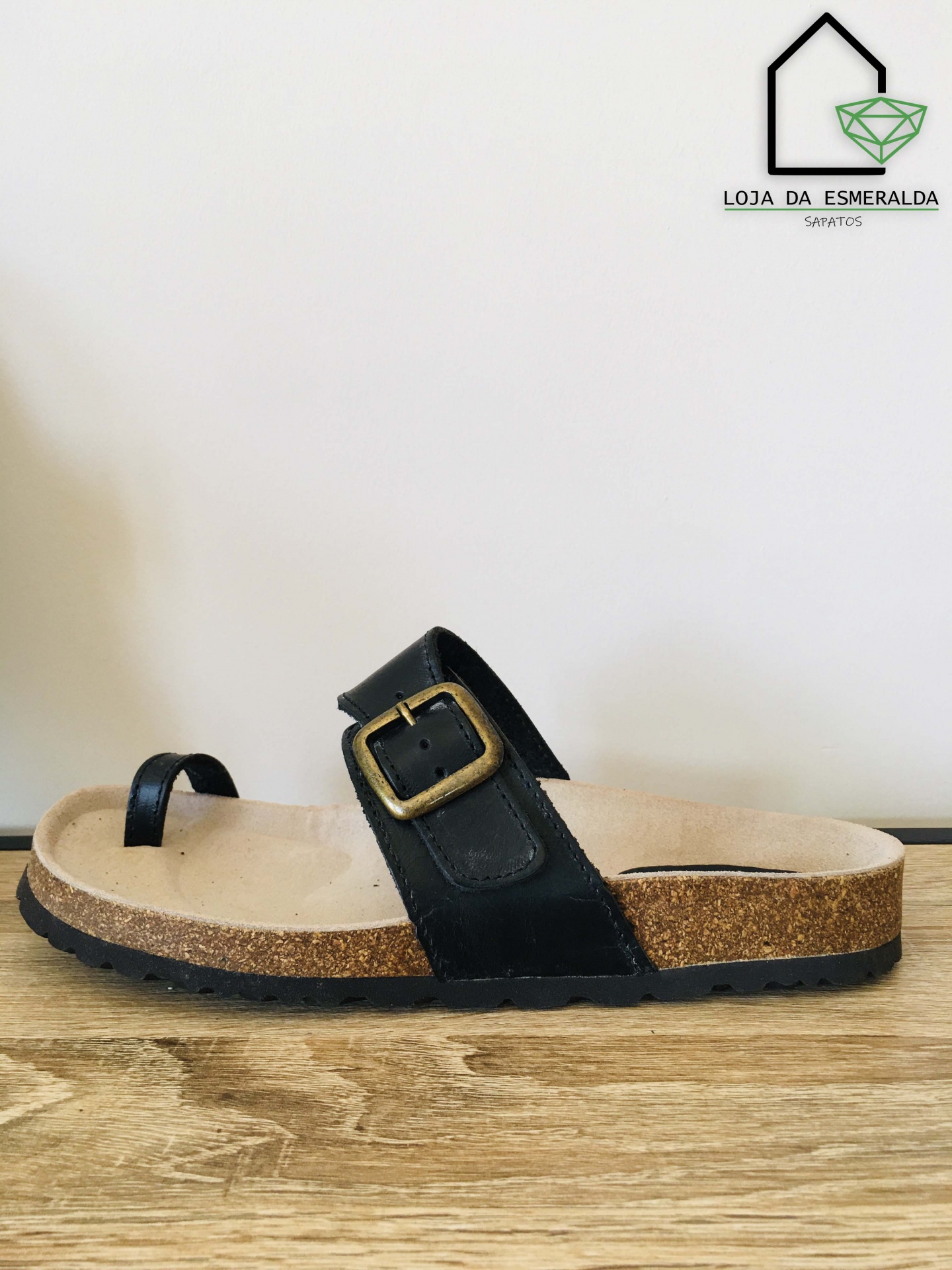 Sandália preta com sola de cortiça em superfície de madeira e logótipo da loja ao fundo