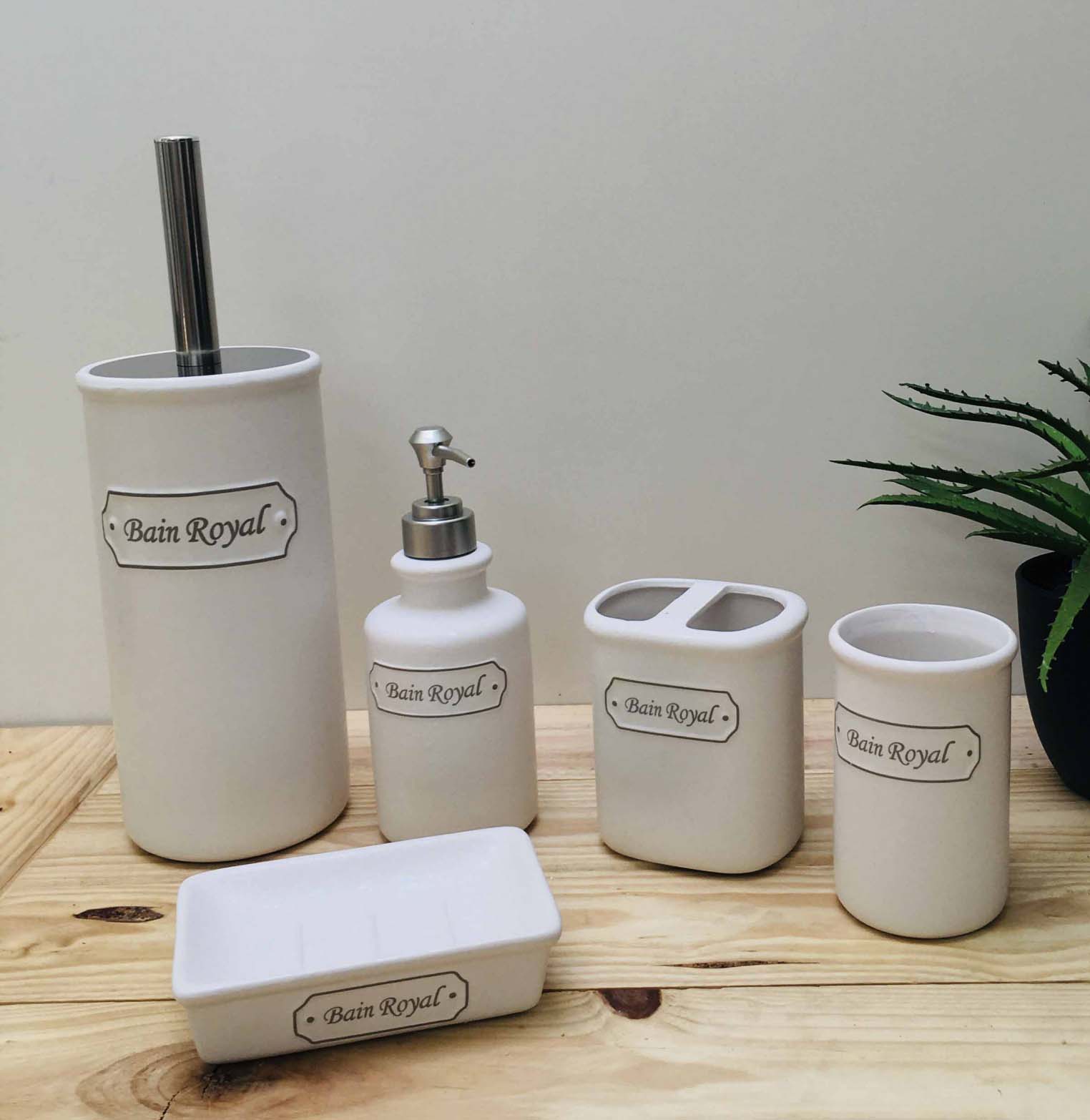 Conjunto de acessórios de casa de banho em cerâmica branca com etiqueta 'Bain Royal' sobre mesa de madeira clara