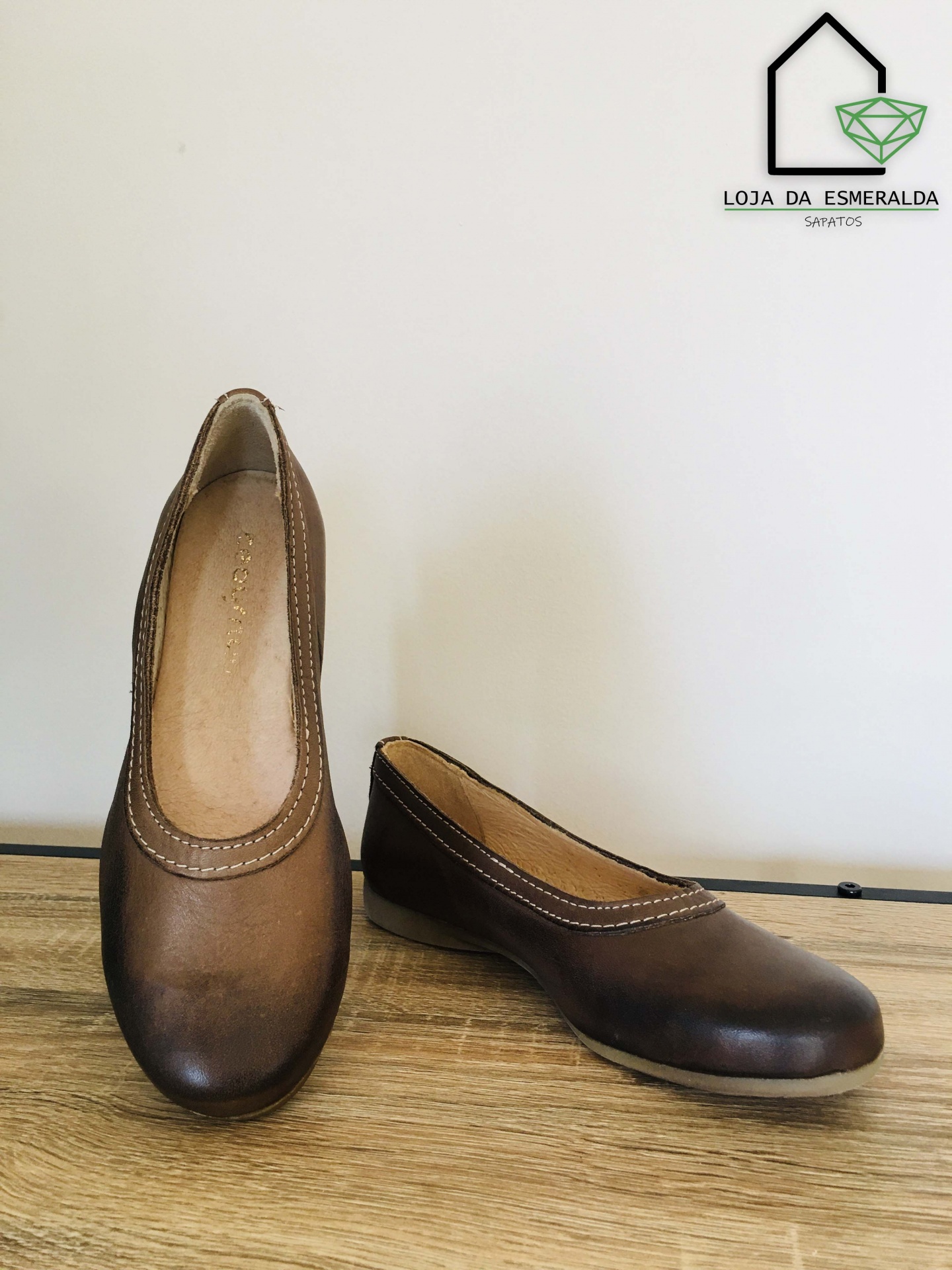 Sapatos tipo sapatilha em pele castanha com costuras brancas sobre superfície de madeira