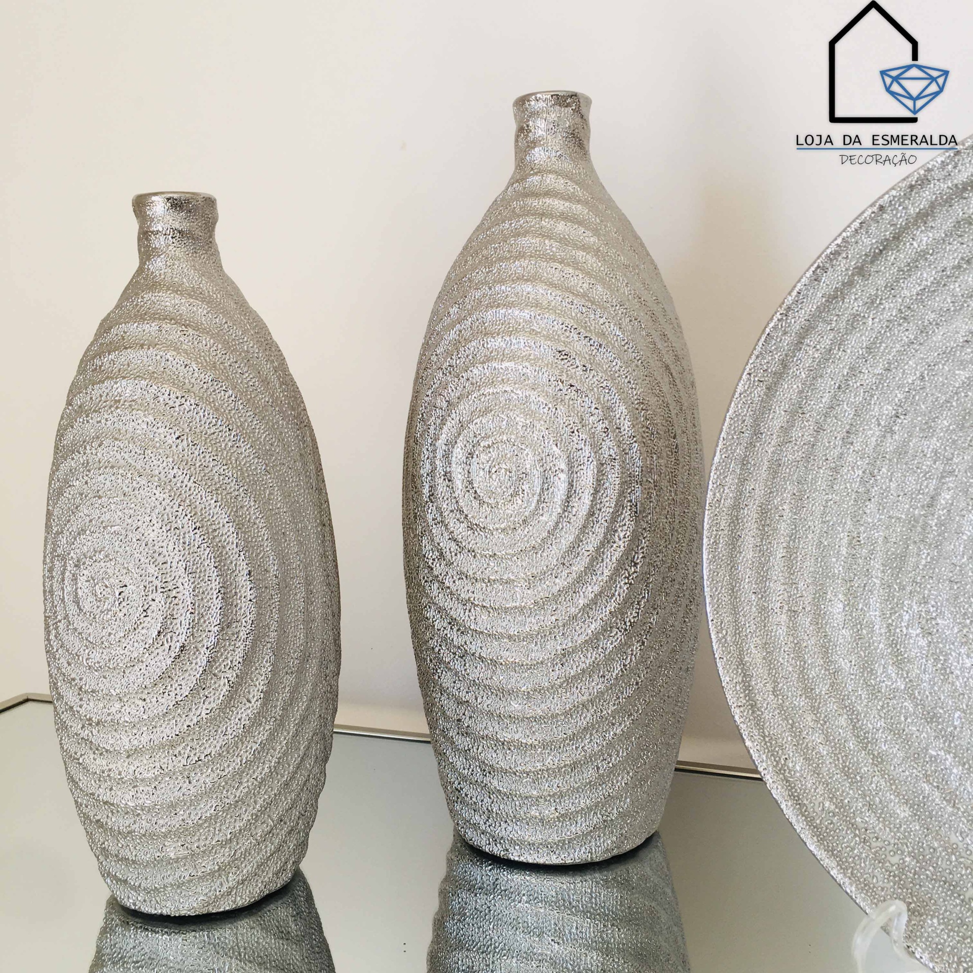 Dois vasos decorativos prateados com padrão circular texturizado sobre superfície espelhada, logo LOJA DA ESMERALDA
