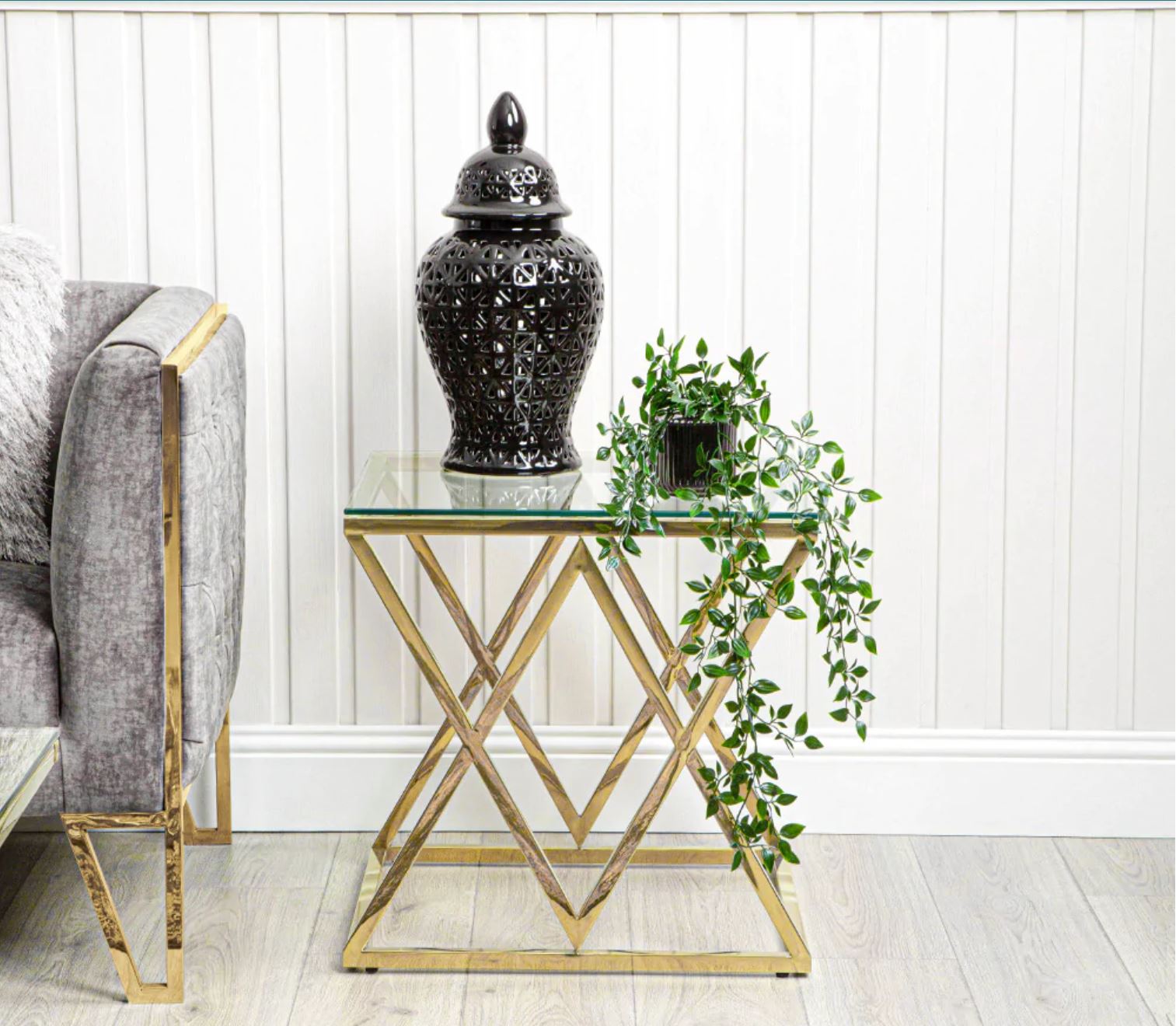 Mesa lateral dourada com tampo de vidro, vaso preto decorativo e planta verde ao lado de sofá cinza