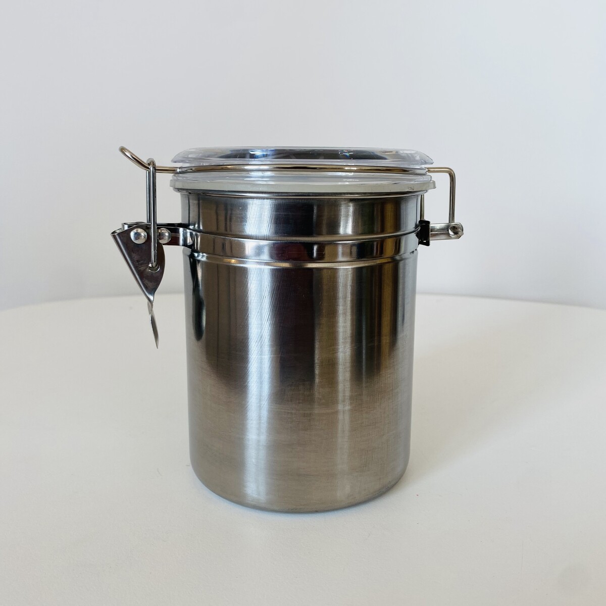 Recipiente cilíndrico de metal prateado com tampa transparente e fecho metálico em fundo branco