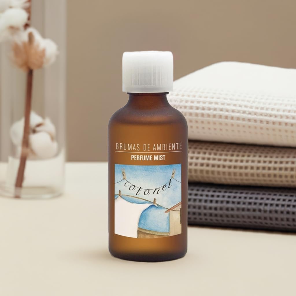 Frasco âmbar de brumas de ambiente com etiqueta azul e branca sobre superfície bege com tecidos dobrados ao fundo