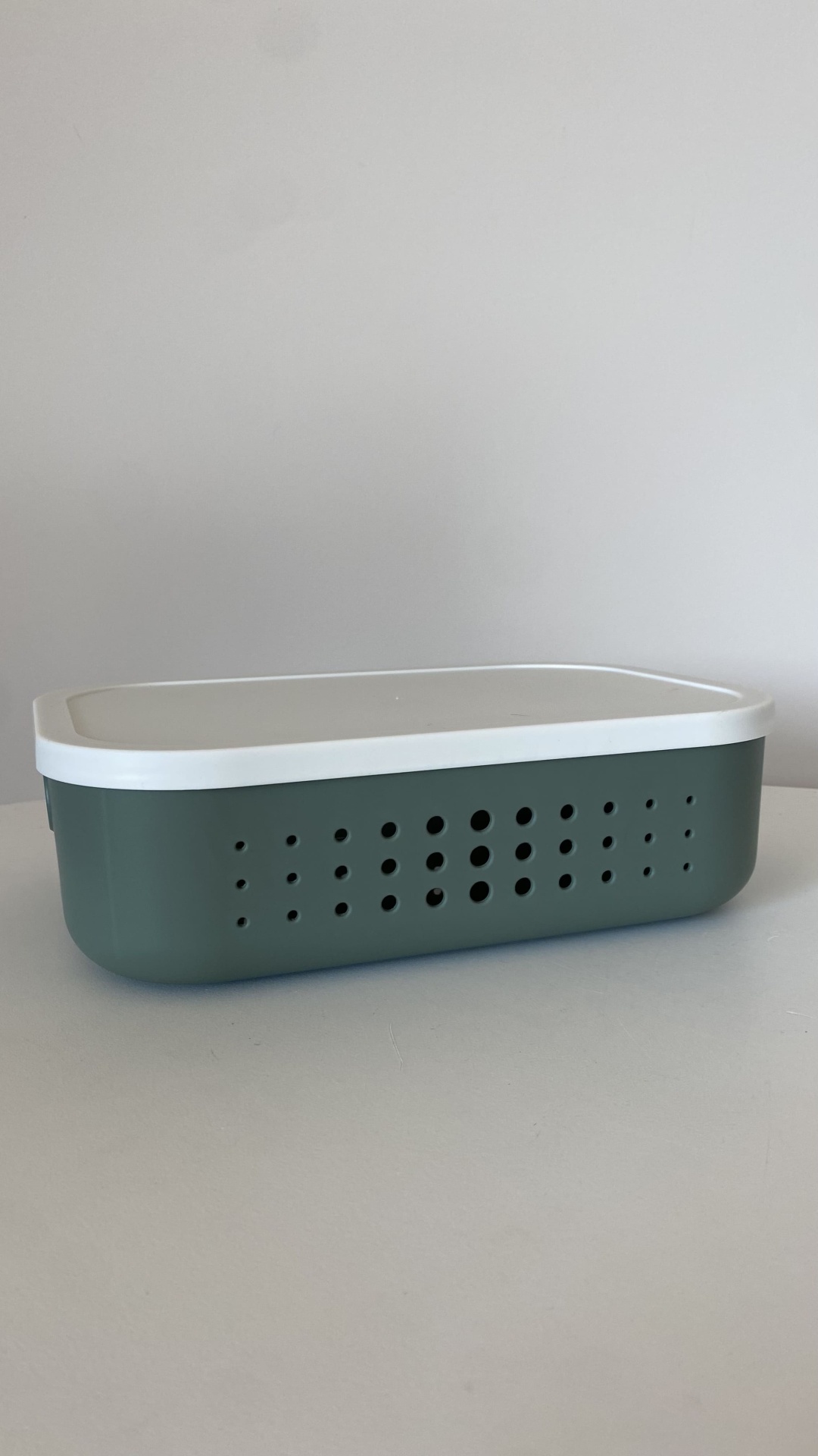 Caixa de plástico verde com tampa branca e orifícios circulares sobre superfície e fundo neutros