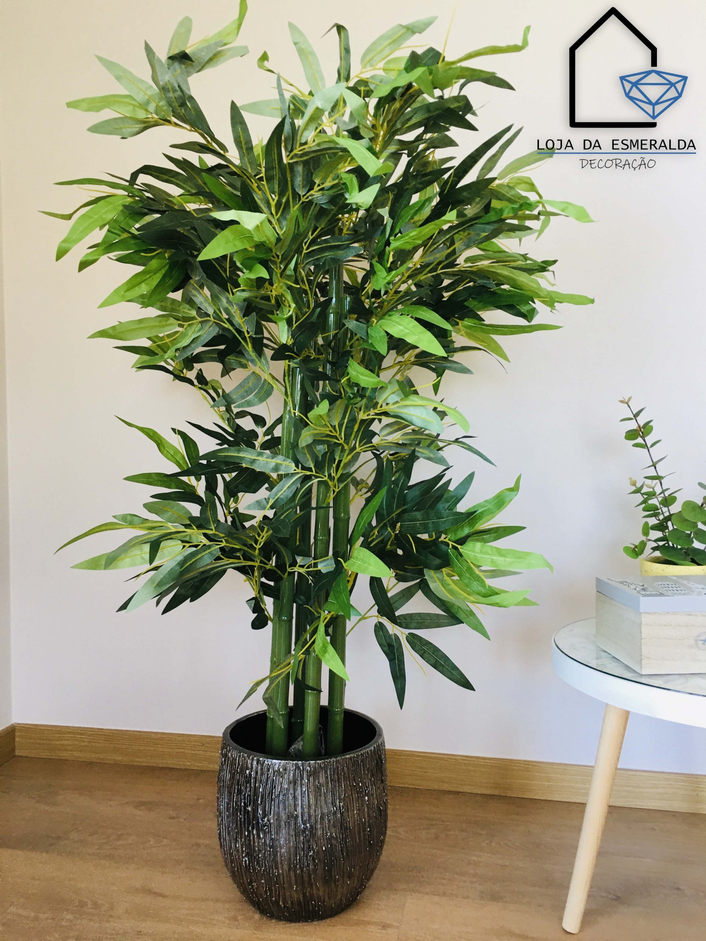 Planta artificial de bambu verde em vaso cinza com texturas, ambiente interior com chão de madeira clara