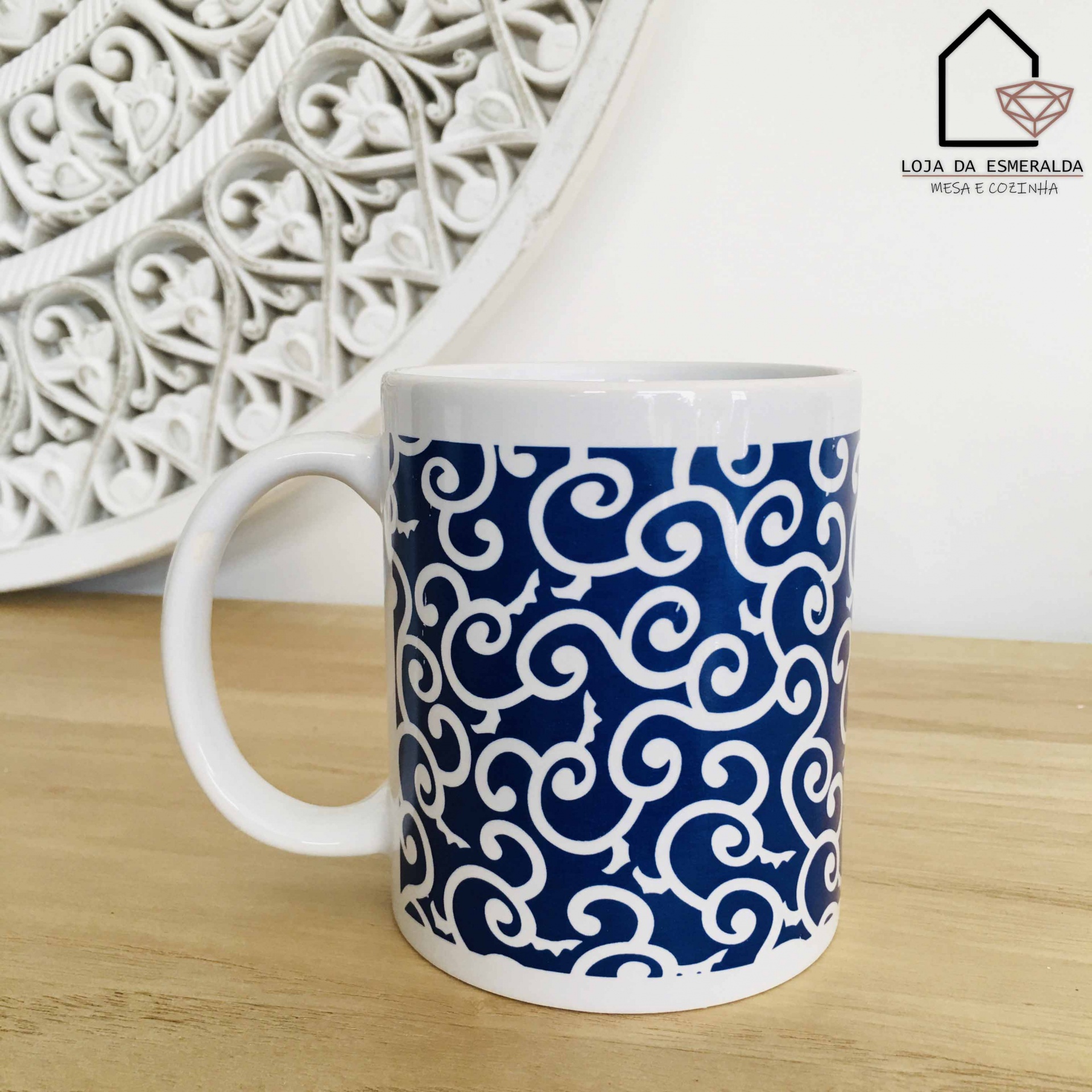 Caneca branca com padrão azul e branco com arabescos, sobre mesa de madeira clara com fundo decorativo branco.
