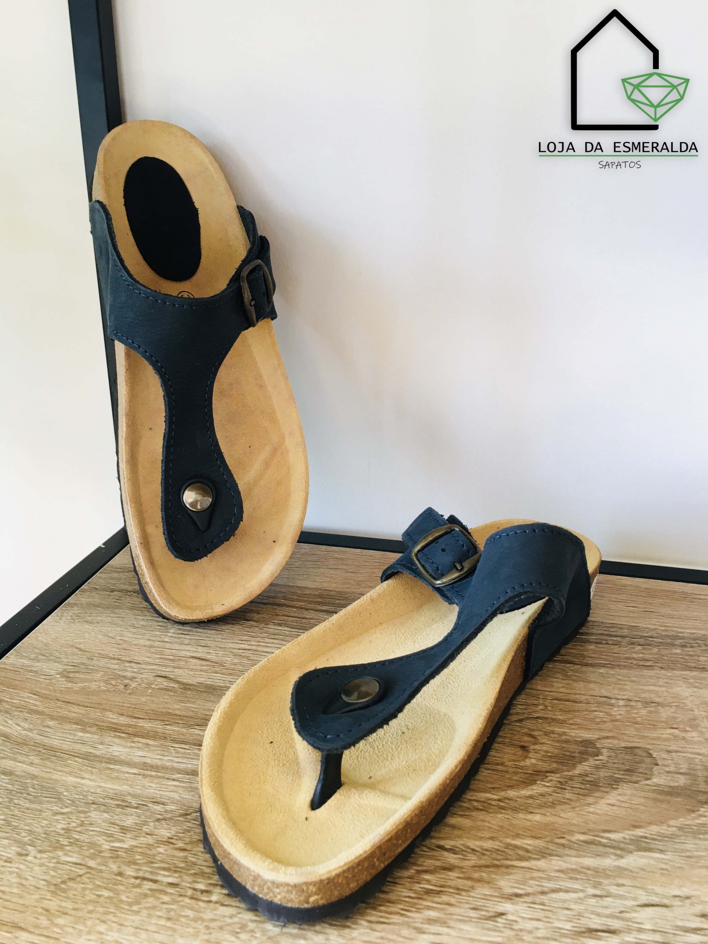 Sandálias de dedo pretas com sola castanha clara em superfície de madeira e logótipo de loja ao fundo