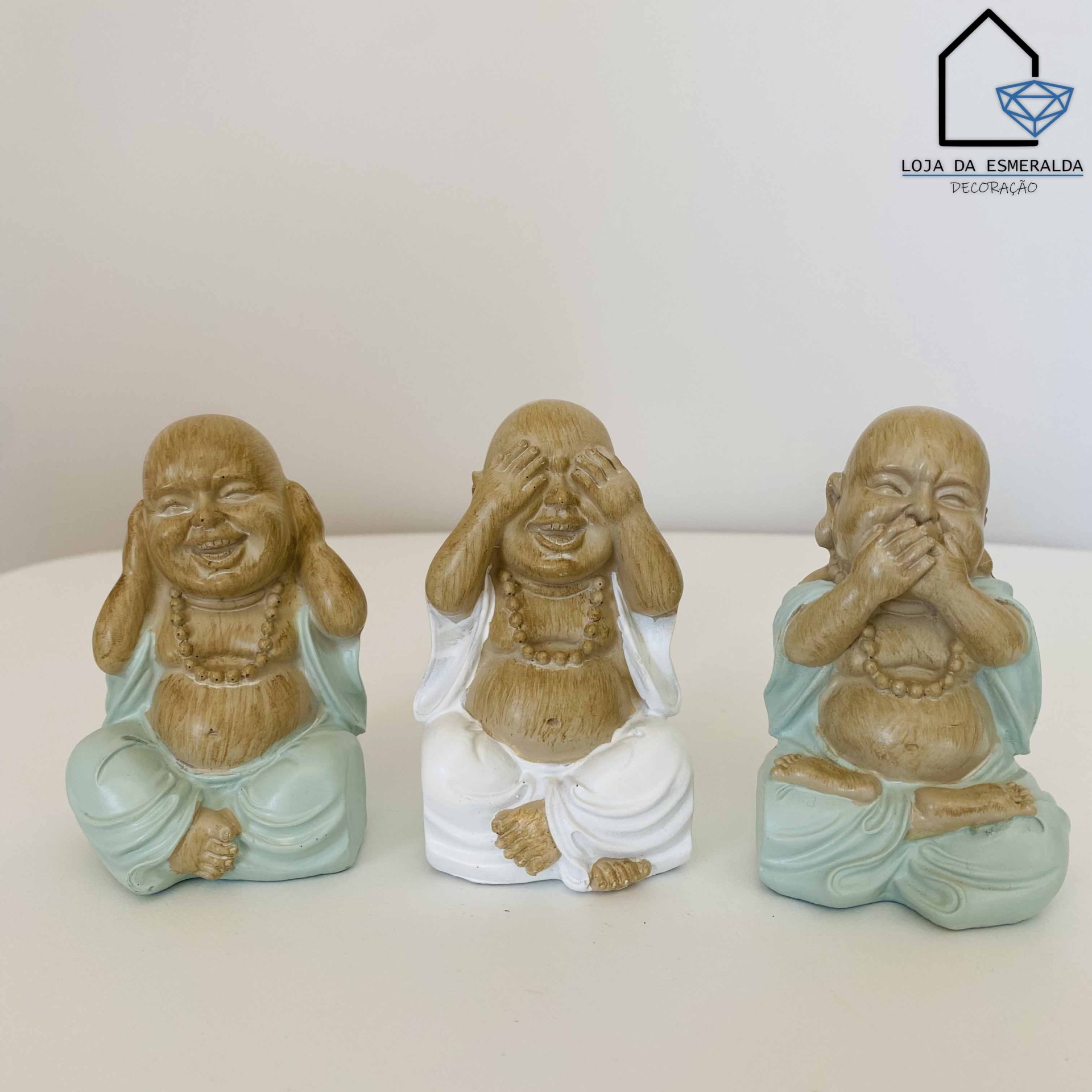 Três estatuetas de Buda sentadas com vestes verdes e brancas, mostrando gestos de cobrir olhos, ouvidos e boca.